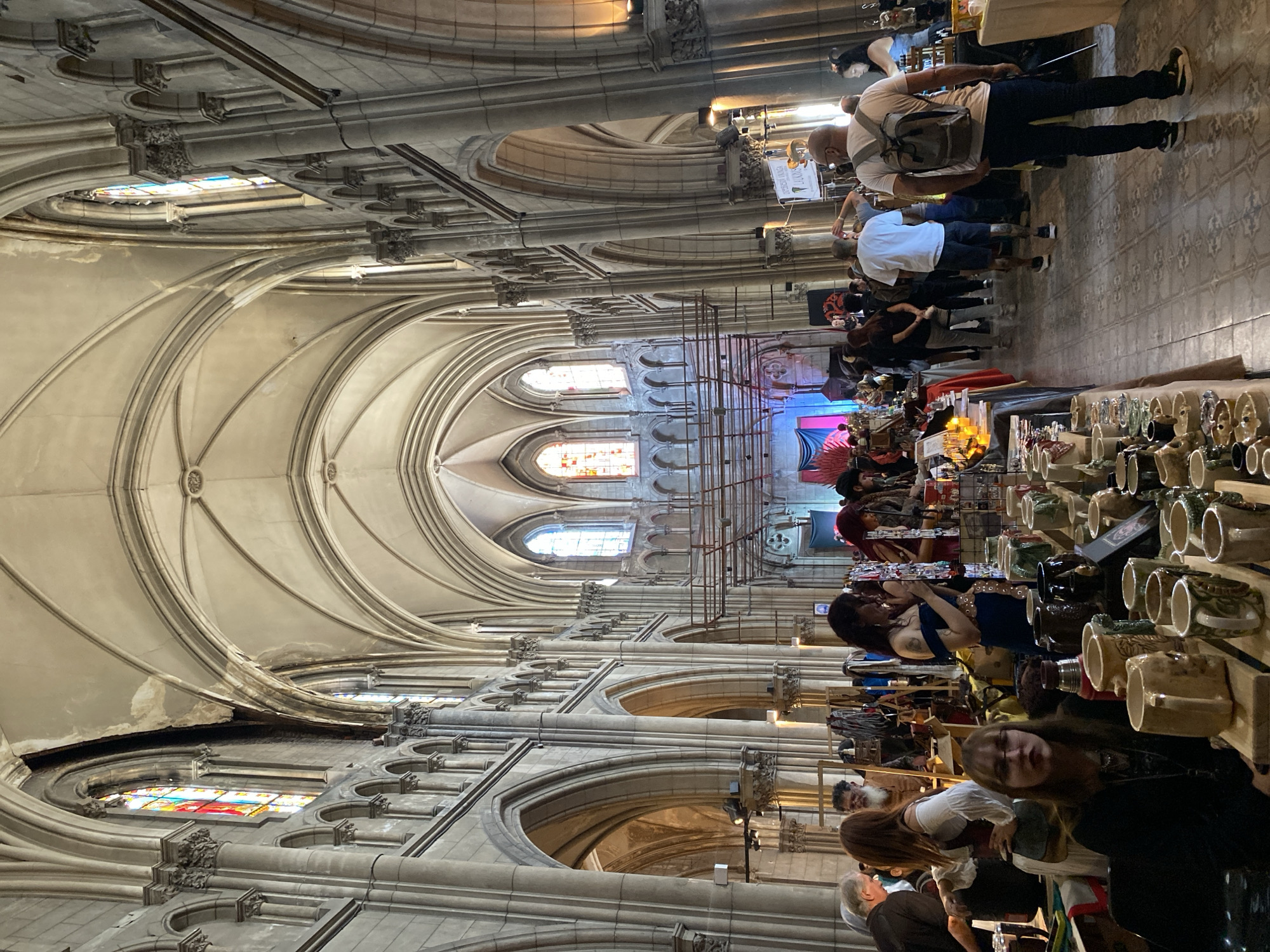 The image shows an indoor market scene, possibly a craft fair, set within a cathedral-like structure with high vaulted ceilings, arches, and stained-glass windows. Numerous vendors have set up tables displaying their wares, ranging from pottery to various artisanal crafts. People of various ages and appearances are browsing the booths, some interacting with the vendors. The architecture suggests a European Gothic or Neo-Gothic style. The image's location within Buenos Aires, Argentina, cannot be verified from visual evidence alone.