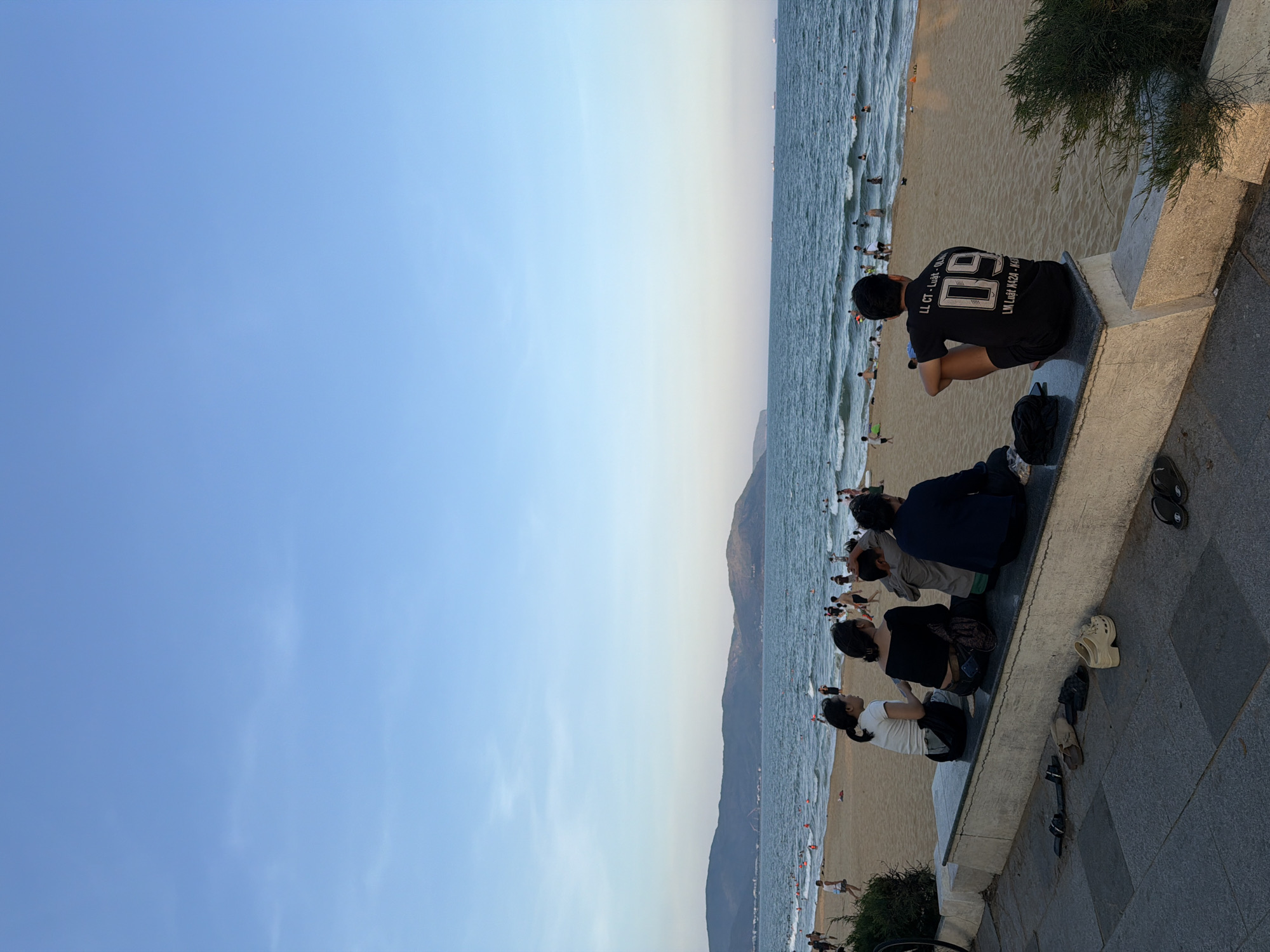 A group of five people are sitting on a low stone wall overlooking a beach and the ocean. The people on the wall are facing away from the camera, towards the water. The person on the far right is wearing a black t-shirt with the number "09" on the back. The person next to them, to their left, is wearing a dark blue long-sleeved shirt. The person to their left is wearing a grey hooded shirt. The person next to them is wearing a black off-the-shoulder top. The person on the far left is wearing a white short-sleeved shirt. There are several pairs of shoes scattered on the ground in front of the wall. The beach behind them is populated with many people, some walking and others in the water. The ocean is a deep blue and appears to have small waves. In the distance, there are mountains that form a coastline. The sky is a clear, pale blue with a few wispy clouds. The scene appears to be in the late afternoon or early evening, judging by the lighting. The location is the coast of Quy Nhon Ward, Vietnam.