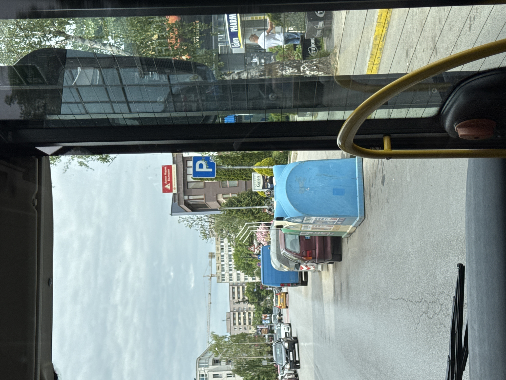 The image is taken from inside a bus or public transport vehicle, looking out the window towards a city street. The view shows a line of parked cars along the right side of the street, with buildings visible in the background. There is a blue bus shelter or structure next to the parked cars. A parking sign with a blue "P" is visible near a building. Further in the distance, the scene shows buildings with a construction crane, indicating ongoing construction or development. Reflections in the glass show a pharmacy and a person outside. The location is Sofia, Bulgaria.