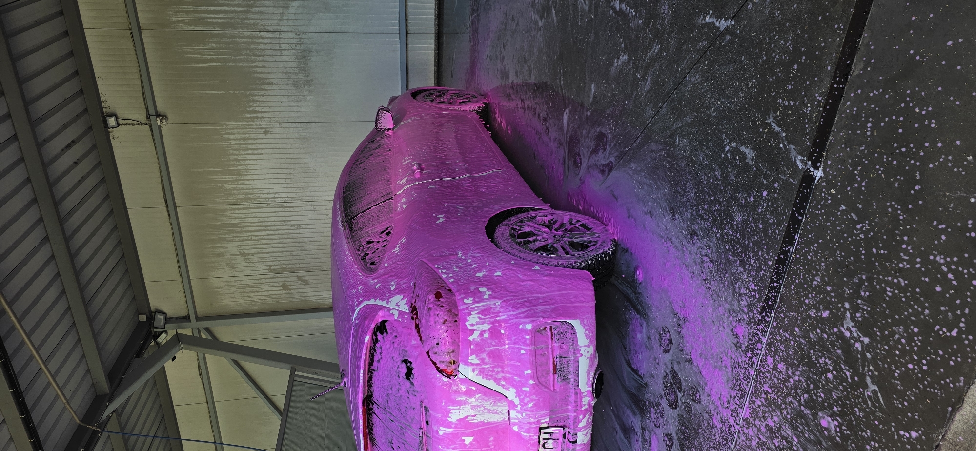 The image depicts a car covered in pink foam in what appears to be a car wash bay. The car, a hatchback, is positioned on a wet, dark surface that reflects the pink foam and the ambient light. The car wash bay has a corrugated metal roof supported by visible metal beams and a ribbed metal wall in the background. There are lights mounted on the ceiling structure. The ground is wet and covered with foamy residue, indicating recent washing activity. The license plate of the car is partially visible and appears to read "CH".
