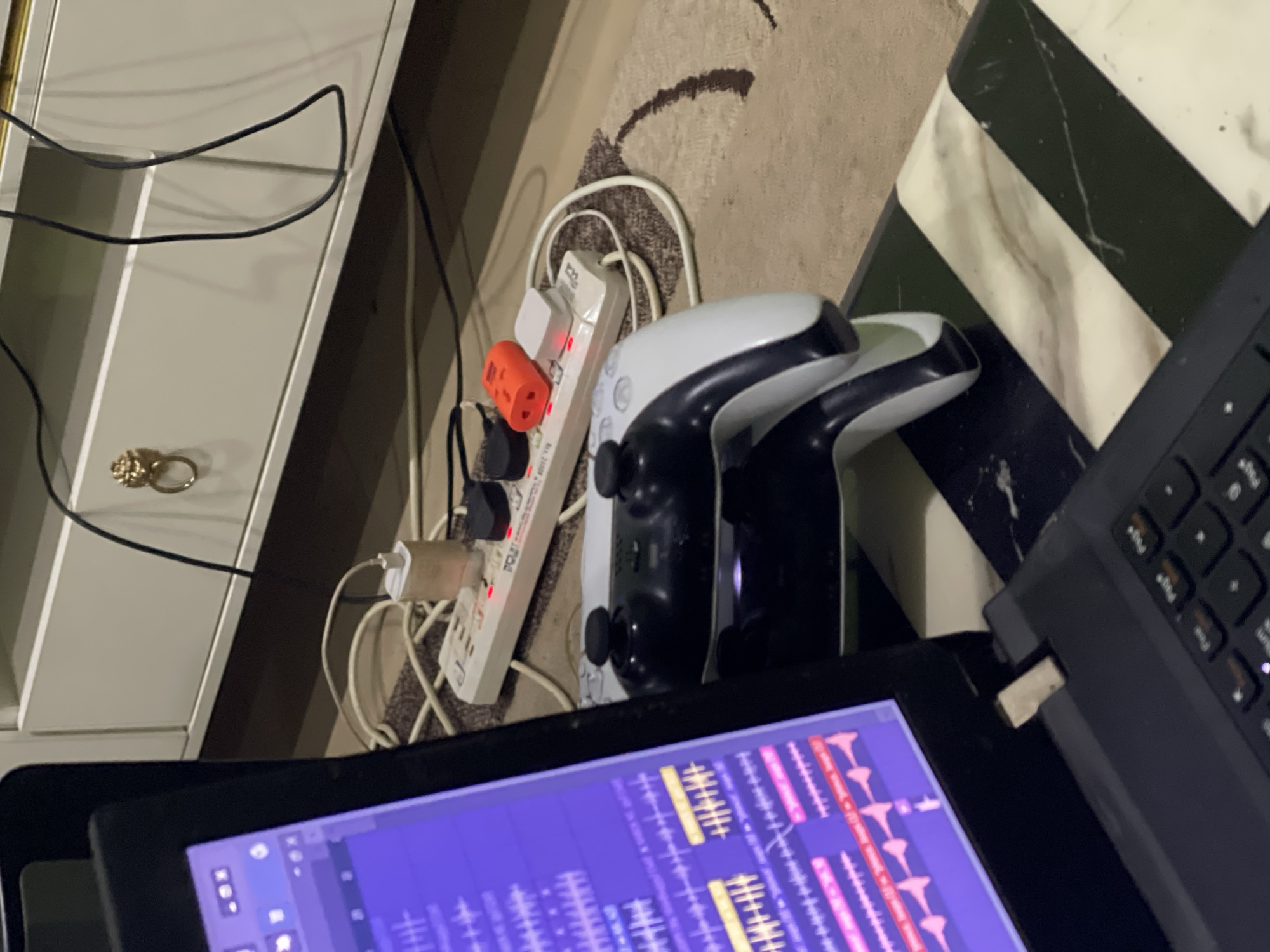 A slightly angled, medium close-up shot depicts an indoor setting. A white power strip with multiple plugs is plugged into a wall outlet. Connected to the power strip are various adapters and a bright orange power adapter. Two white Playstation 5 controllers rest next to the power strip, positioned at a slight angle. In the foreground, a black laptop screen displays a music production software interface with numerous waveform patterns and editing tools. To the right, part of a laptop keyboard is visible. The background is a mottled beige and brown rug. The overall lighting of the scene is somewhat dim, with a warm color cast. The scene is set on a surface with a black and white marble pattern. The image does not offer sufficient detail to verify the location, Abuja, Nigeria. There are no people present in the image.