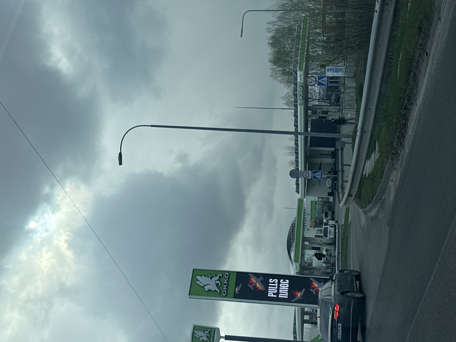 The image shows a gas station in Chabany, Ukraine. It is an overcast day with a gray sky and a few clouds. The gas station is an OKKO brand and is located on a highway. There are two people in the scene, one standing on the sidewalk near the gas station, the other driving a black car. The car is driving toward the viewer. There is a sign on the side of the gas station that reads "Pulls Plus" in Ukrainian. The sign has a red background and depicts three airplanes. There is a tall streetlight with a single light bulb on the left of the scene. There are two additional streetlights with the same design in the distance on the right of the scene. There are also various other cars parked on the lot of the gas station. There are green bushes and trees on the right side of the image, behind the gas station. There is a concrete barrier along the side of the highway, which curves toward the gas station on the left of the image.
