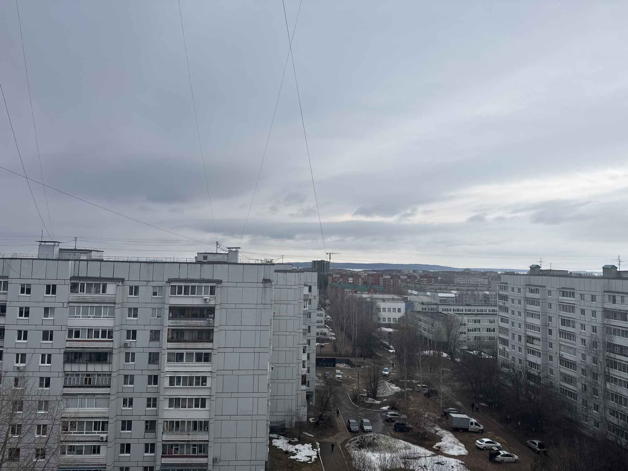 An elevated perspective of a residential area in the city of Chaikovsky, Russia, on an overcast day. In the foreground, two grey concrete apartment buildings flank a view of a street below. The street is unpaved in sections and exhibits patches of melting snow along the edges. A few parked cars and a white van are visible on the street. Bare trees line the street.
In the distance, a cityscape is discernible, featuring other residential buildings and a skyline marked by a tall construction crane. The sky is filled with grey clouds. Electrical wires crisscross the upper portion of the image.