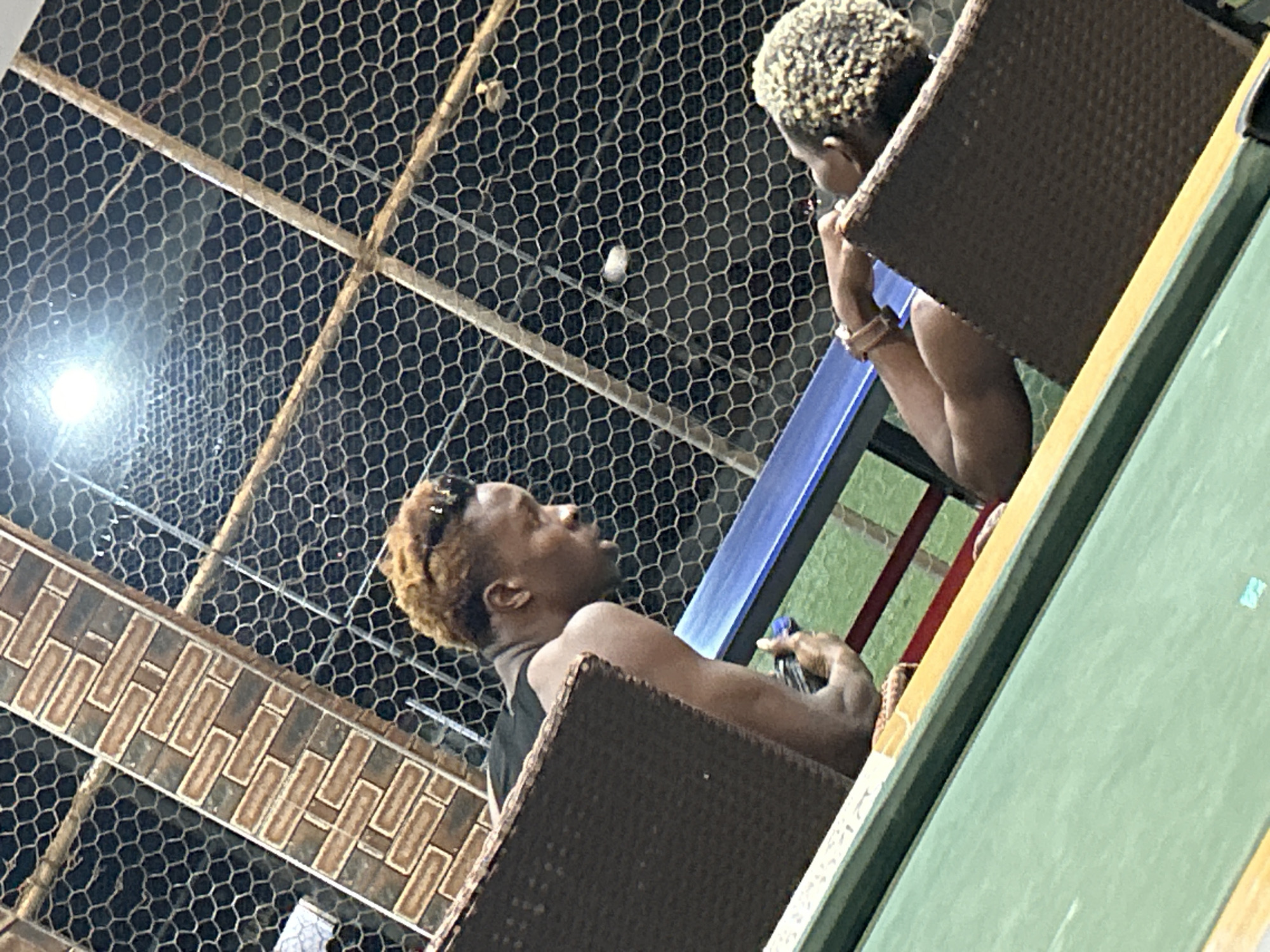 Two individuals are seated at tables, with a mesh fence in the background. One individual, with light brown hair, is leaning forward, looking down and to the right. They are wearing a black tank top and their left arm is resting on a table. The second individual, with tightly curled blonde hair, is visible from the back and is also leaning forward, with their right arm resting on their lap and their left arm extended. A small white object is visible in the mesh in the background. The setting appears to be outdoors.
