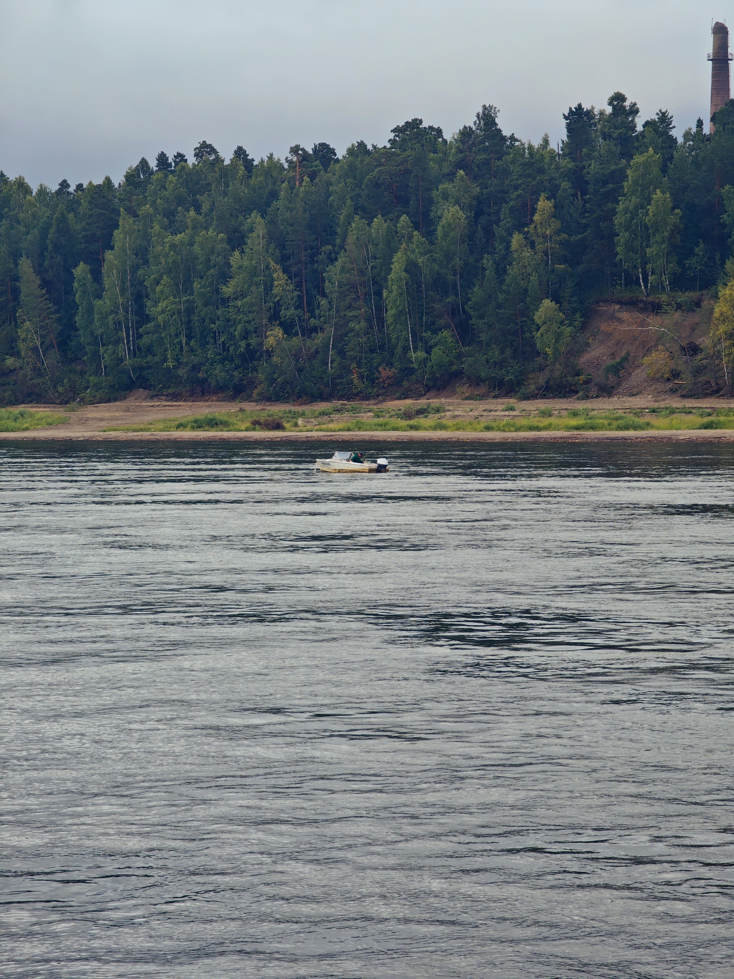 A small, yellow boat with a white canopy and an outboard motor is on a wide, dark river under an overcast sky. The boat is located in the mid-ground of the image, positioned slightly to the left of center. Behind the river, a steep, wooded embankment rises, covered in a dense forest of green deciduous and coniferous trees. The embankment has a sandy, eroded slope visible in places. In the upper right corner of the image, a tall, brick smokestack with a metal platform near its top rises above the trees, indicating industrial activity. The river's surface is rippled, reflecting the muted light. The scene appears to be set in a natural, possibly rural or industrial area bordering a river. The image was taken in the city of Lesosibirsk, Russia.