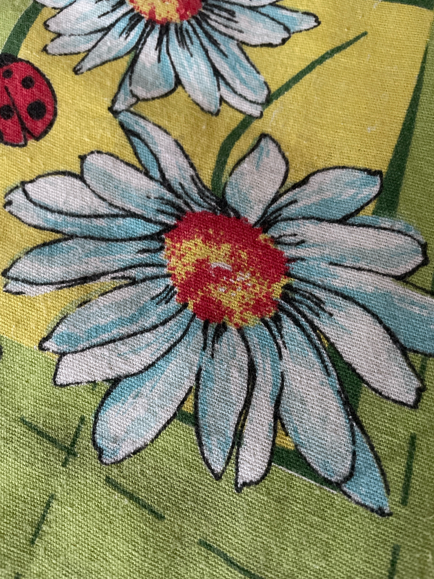The image shows a close-up, overhead view of a fabric with a floral pattern. The pattern features a large daisy with light blue and white petals outlined in black, and a red and yellow center. To the left of the daisy, there is a small red ladybug with black spots. The background is a yellow square with green lines, and the outer edges of the fabric are green. The fabric appears to be a textured cotton or linen material. The scene is indoors and the lighting is natural and even.