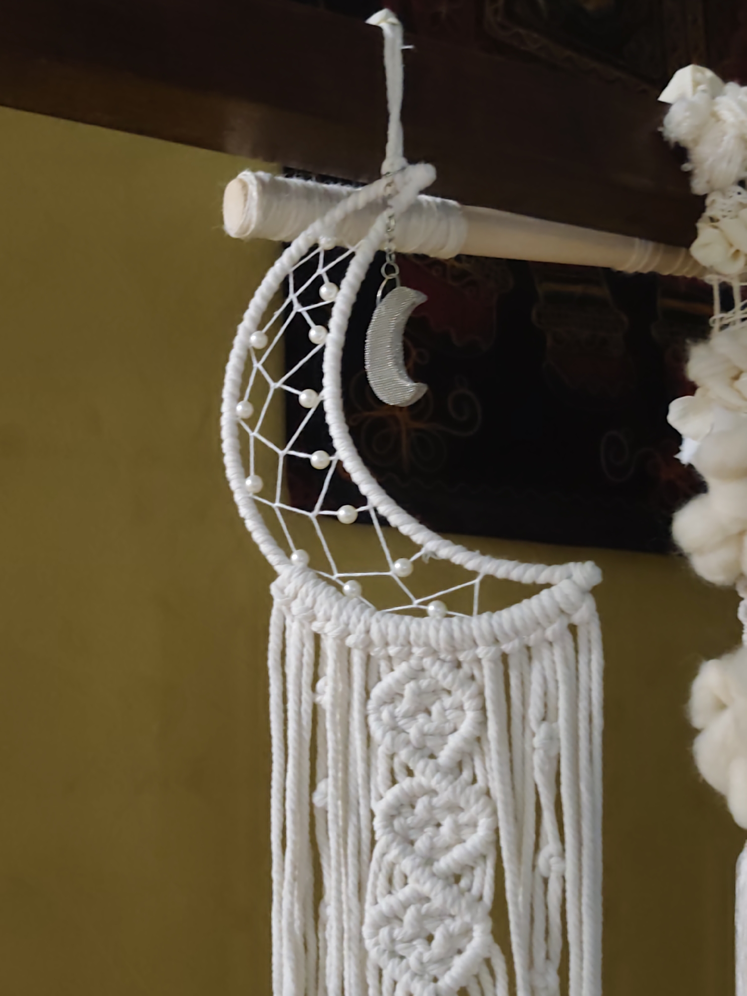 This is a close-up, vertical, medium shot of a white macrame dreamcatcher hanging from a wooden beam. The dreamcatcher has a crescent moon shape and is adorned with white pearls. Below the crescent, macrame cords hang down and are intricately knotted. A decorative, fluffy white object is visible on the right side of the frame, partially obscured. The background consists of a textured mustard-yellow wall. The scene appears to be indoors, possibly in a residential setting. No specific landmarks of Sochi, Russia, are visible.