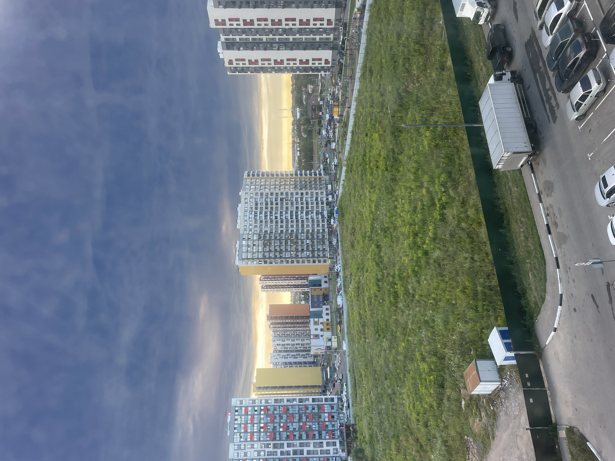 The image depicts a city scene in Domodedovo, Russia, characterized by a mix of residential buildings, green spaces, and infrastructure. In the foreground, a paved area with white and black curbs shows several parked cars, including a silver sedan, a dark sedan, and a white car. A large white truck with a box trailer is also parked on the paved surface. Adjacent to this is a green fence, behind which lies a field of tall green grass, interspersed with some yellow wildflowers. Two small utility structures, one white with blue doors and another with a rusted roof, are visible within the grassy area.
Beyond the grassy field, a row of multi-story residential buildings dominates the middle ground. These buildings vary in color and design, with some featuring colorful balconies or facades. A cluster of smaller, brightly colored buildings that appears to be a school or kindergarten is situated among the residential blocks. Beyond these, more residential buildings and a distant cityscape are visible under a cloudy sky. The sky itself is a dramatic blend of deep blues and grays, with patches of lighter clouds near the horizon, suggesting either sunset or an approaching storm. The overall impression is one of a developing urban area with ample green space, situated in a location identifiable as Domodedovo, Russia.