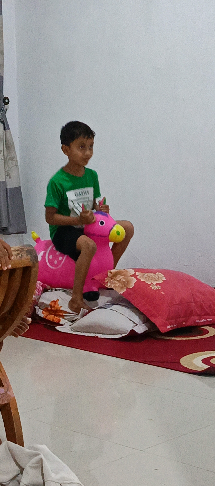 A young boy with short, dark hair and brown eyes is sitting on a bright pink inflatable toy horse. He is wearing a green t-shirt with a white and grey design that appears to say "GAZILE". His legs are bent, and his feet are visible. The horse has yellow eyes and a yellow nose. The boy is holding onto the horse's reins.
The scene is indoors, in what appears to be a room with a light-colored wall. In the foreground, there are several pillows and a red rug. One pillow has a floral pattern in shades of red and beige. The floor is tiled. To the left of the frame, a wooden chair leg is visible, and some light-colored fabric is draped over it.
The overall setting suggests a casual, domestic environment. The image does not provide any specific contextual information that would allow for the identification of the city of Pashiebeutong, Indonesia.