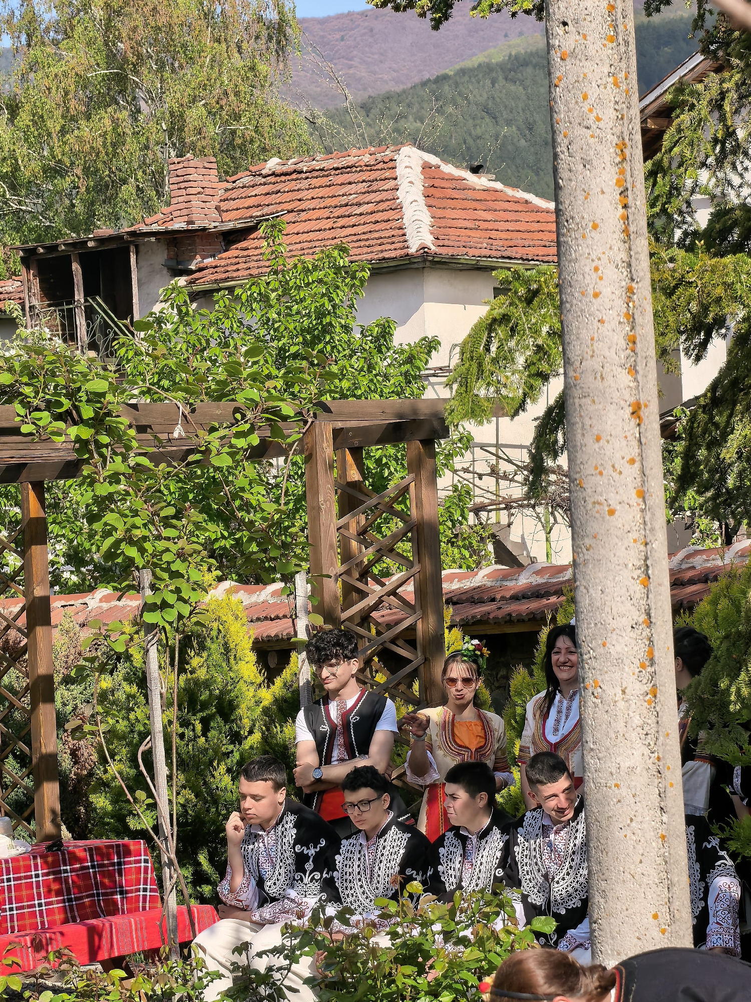 In a sunny outdoor setting, a group of people in traditional Bulgarian clothing are gathered. The location appears to be in Sapareva Banya, Bulgaria, given the architectural style and cultural attire. Several young men are seated on a bench covered with a red plaid cloth, wearing white shirts with ornate black and white embroidery, paired with white pants. Some other individuals, including women, are standing or seated nearby, dressed in similar traditional garments. In the background is a building with a red tiled roof, and a wooden pergola covered in foliage. A concrete pillar covered in orange lichen stands in the foreground. The mood appears relaxed and celebratory, possibly a cultural event or gathering. Hills or mountains are visible in the distance.