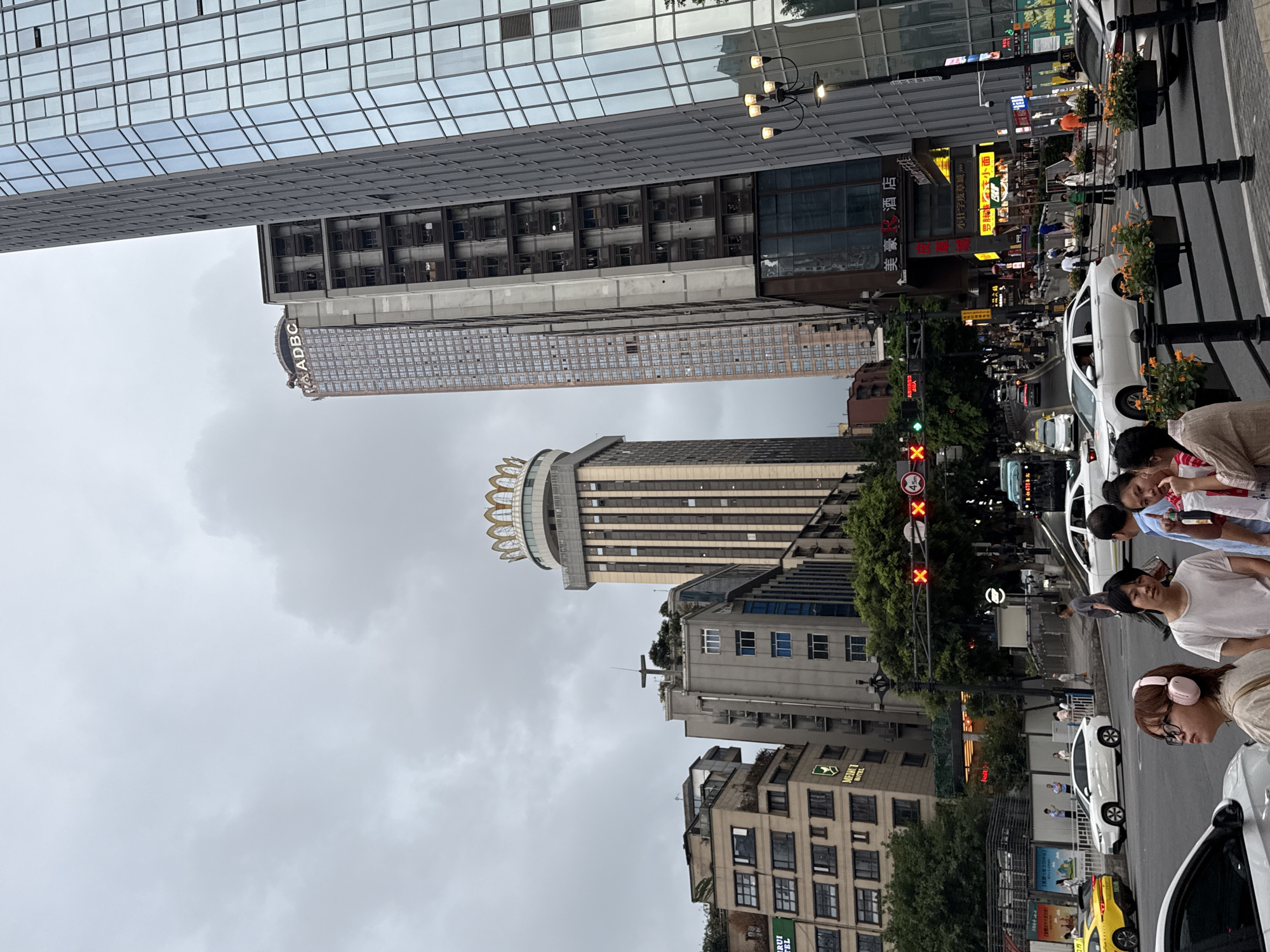 The image depicts a street scene in Yuzhong District, China, under an overcast sky. Tall modern buildings dominate the skyline, some with glass facades reflecting the surroundings. In the foreground, a street with cars and pedestrians is visible. A traffic light displays a red signal. Several people are standing on the sidewalk, some appearing to be waiting or walking. Plants in planters add a touch of greenery to the urban landscape. The overall impression is that of a bustling city environment on a cloudy day.