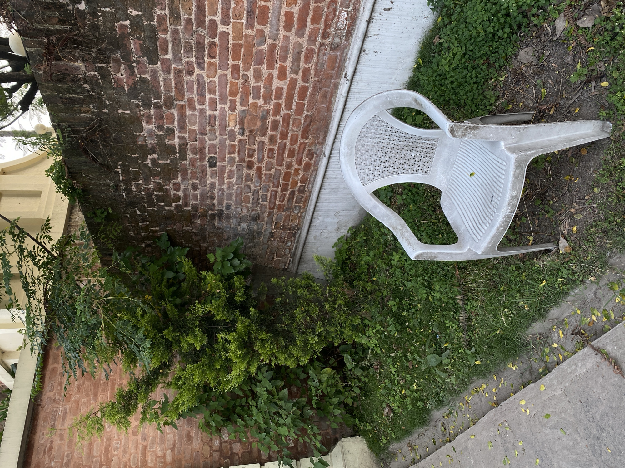 An outdoor scene in Kathmandu Metropolitan City, Nepal, features a white plastic chair placed on grass next to a brick wall and a concrete path. The chair is positioned in the foreground, angled slightly to the right, revealing its worn and textured surface. Behind the chair, a lush green bush grows against the red brick wall, which shows signs of age and weathering. A concrete channel runs along the edge of the path, filled with fallen yellow and green leaves. The environment suggests a somewhat neglected urban outdoor space, possibly a garden or a back alley.