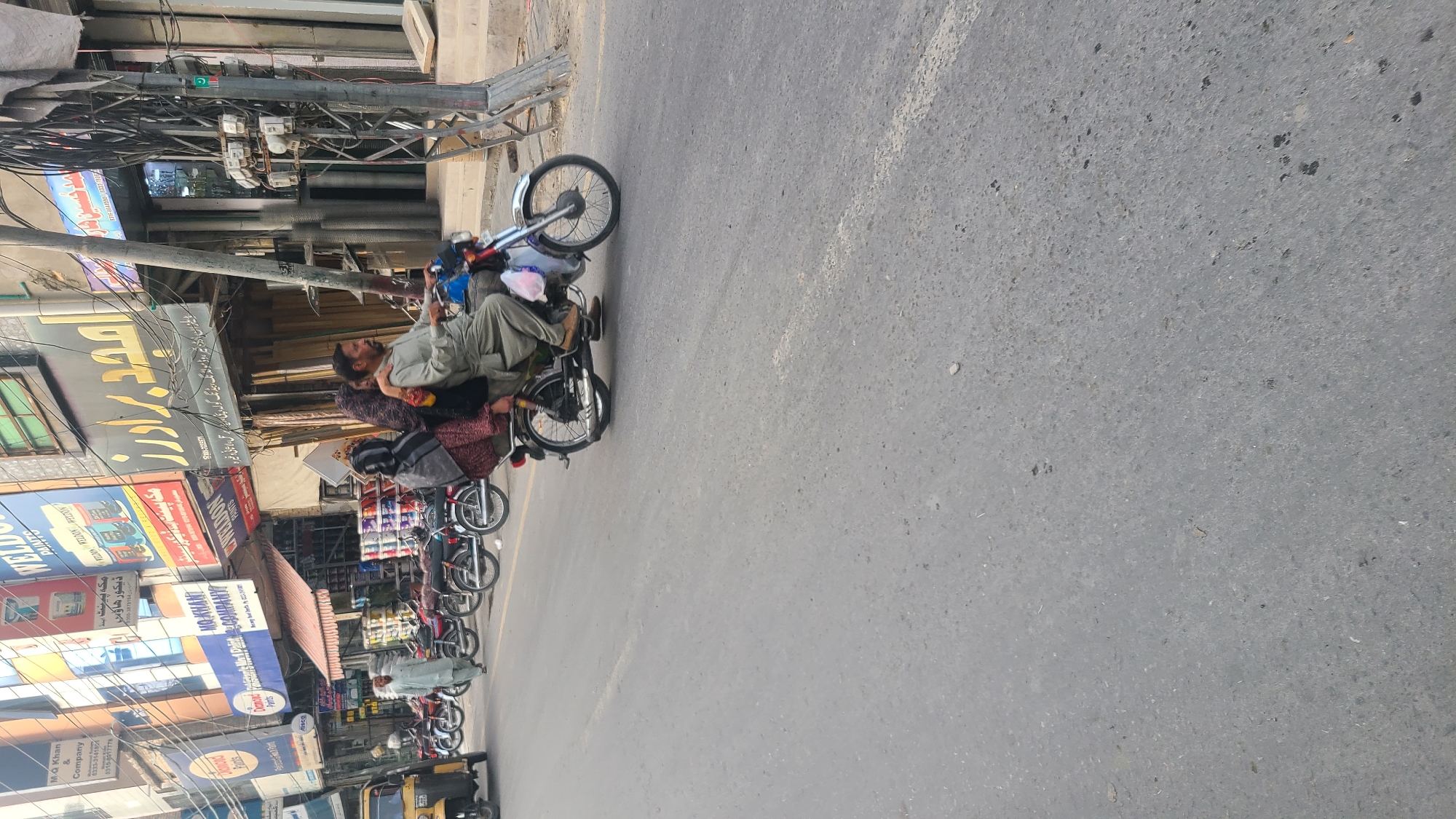 The image shows a man reclined and sleeping on a parked motorbike along a city street in Quetta City Tehsil, Pakistan. Other parked motorbikes line the curb behind the first. Buildings with shopfronts are visible, as are overhead power lines. Signage is present in Urdu and English, including the name "MQ Khan & Company."