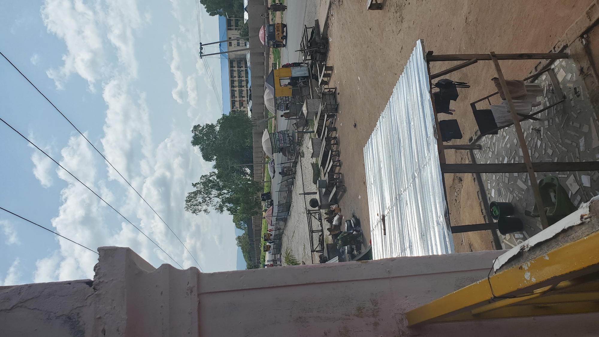 The image shows an outdoor scene in what appears to be a market or roadside area. In the foreground, a partially visible structure with a corrugated metal roof is prominent, supported by wooden beams. Below this structure, a tiled floor is visible, with two black buckets and a green basin containing water. Clothing is hanging on a drying rack under the shelter.
Further back, a road runs horizontally across the image, lined with various stalls and vendors. Some stalls have umbrellas providing shade, and others appear to be small kiosks. Several three-wheeled vehicles, possibly auto-rickshaws, are parked or in motion on the road. People are seen walking and interacting around the stalls.
In the middle ground, there are wooden benches and tables, suggesting an area for seating or display. Further in the distance, more trees are visible, and beyond them, a large building with a blue roof and multiple windows stands out, possibly a school or dormitory. The sky is partly cloudy with a bright, clear blue sky visible between the clouds. The overall impression is of a bustling, everyday scene in a community.