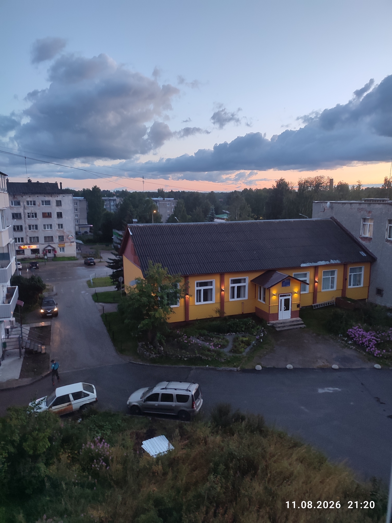 The image captures an aerial view of a residential area in Suoyarvi, Russia, at dusk. The sky is a twilight blue with dramatic clouds tinged with orange and pink near the horizon. In the foreground, a grey SUV is parked on a wet asphalt road. To its left, a white car with a rusted rear section is also parked. A person wearing a blue shirt and dark pants is visible near the white car, appearing to walk away from it.
Dominating the mid-ground is a low, brightly lit yellow building with a red trim and a dark, sloped roof. The building has several windows and an entrance with a small porch. Surrounding the yellow building are gardens with blooming pink and purple flowers. Adjacent to the yellow building is a larger, grey concrete structure with multiple windows.
Further in the background, several multi-story apartment buildings and trees form the urban landscape of Suoyarvi. The streetlights are illuminated, casting a warm glow on the wet road surface, suggesting recent rainfall. The overall scene depicts a quiet evening in a Russian provincial town. A date and time stamp "11.08.2026 21:20" is visible in the lower right corner of the image.