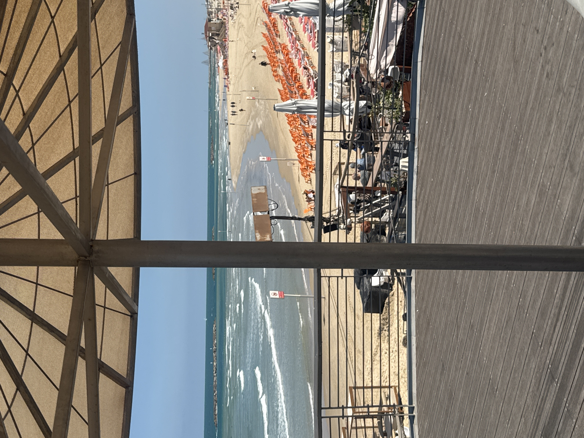 The image depicts a beach scene, likely a portion of Tel Aviv, Israel, captured from an elevated perspective. The foreshore and water meet the coast, with numerous people and sunbathing chairs positioned on the sandy shore. The water presents noticeable wave action and there are stone structures that extend into the sea. A wooden deck runs parallel to the shoreline, delineated by a metal railing. A partial view of a roof structure, presumably from the perspective point, is visible in the lower and left portions of the image. A metal pole is situated horizontally across the center of the image.