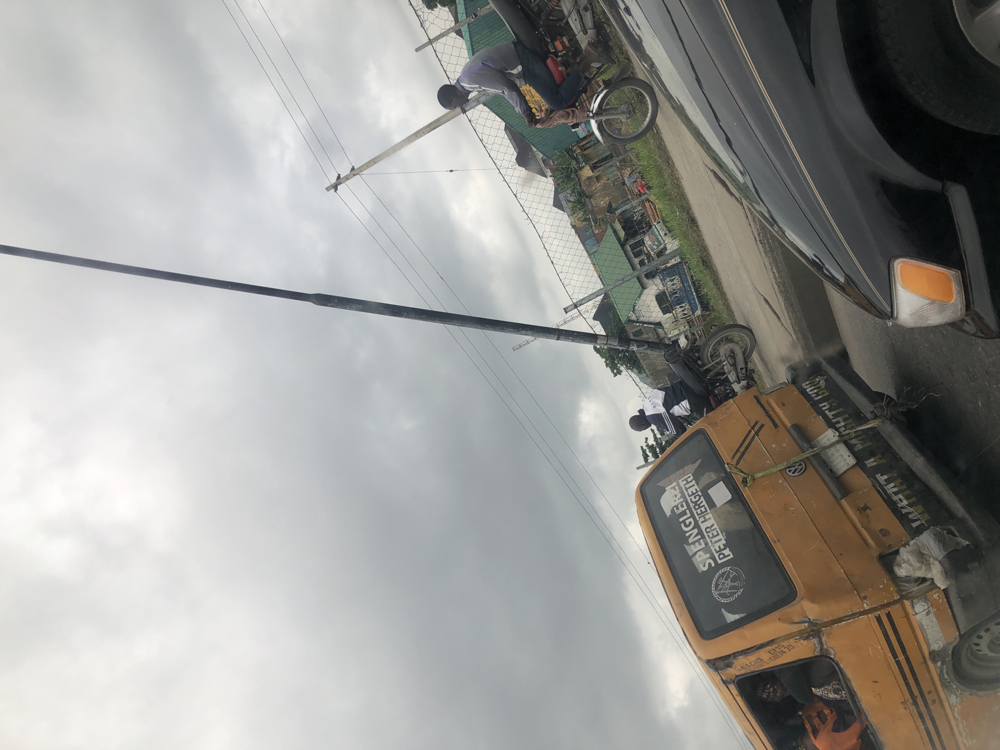 The image captures a street scene in Satellite Town, Nigeria. In the foreground, a black car is partially visible. Behind it, a yellow commercial van with visible damage and graffiti is parked. The van has several stickers on its rear window, one of which reads "SPENGLE PETER HERGETH". Inside the van, passengers are visible. To the right of the van, a man on a motorcycle is riding past. The environment includes a fence, buildings in the background, power lines, and a cloudy sky.