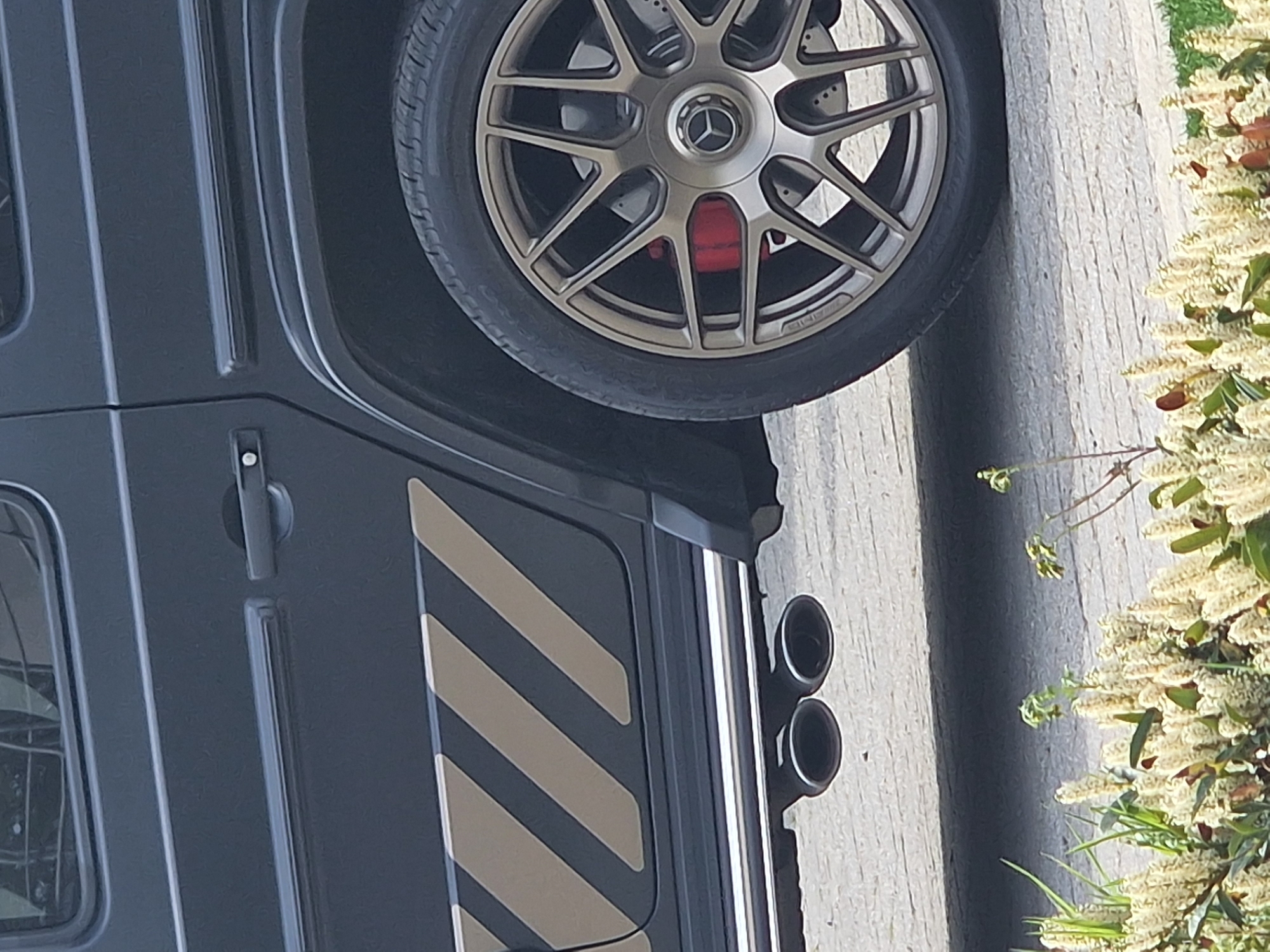 A close-up shot of a dark-colored SUV, presumably a Mercedes-Benz G-Class, is positioned beside a sidewalk in Witton Gilbert, United Kingdom. The SUV features a distinctive wheel with a multi-spoke design and red brake calipers visible behind the wheel. The side of the SUV is adorned with contrasting diagonal stripes and a set of dual exhaust pipes. A shrub with white blossoms is located adjacent to the vehicle. The vehicle and plant are beside a concrete road. The scene is illuminated with soft daylight, indicative of an overcast or diffused lighting condition. No people or other objects are visible. The image is rotated clockwise to orient the landscape vertically.
