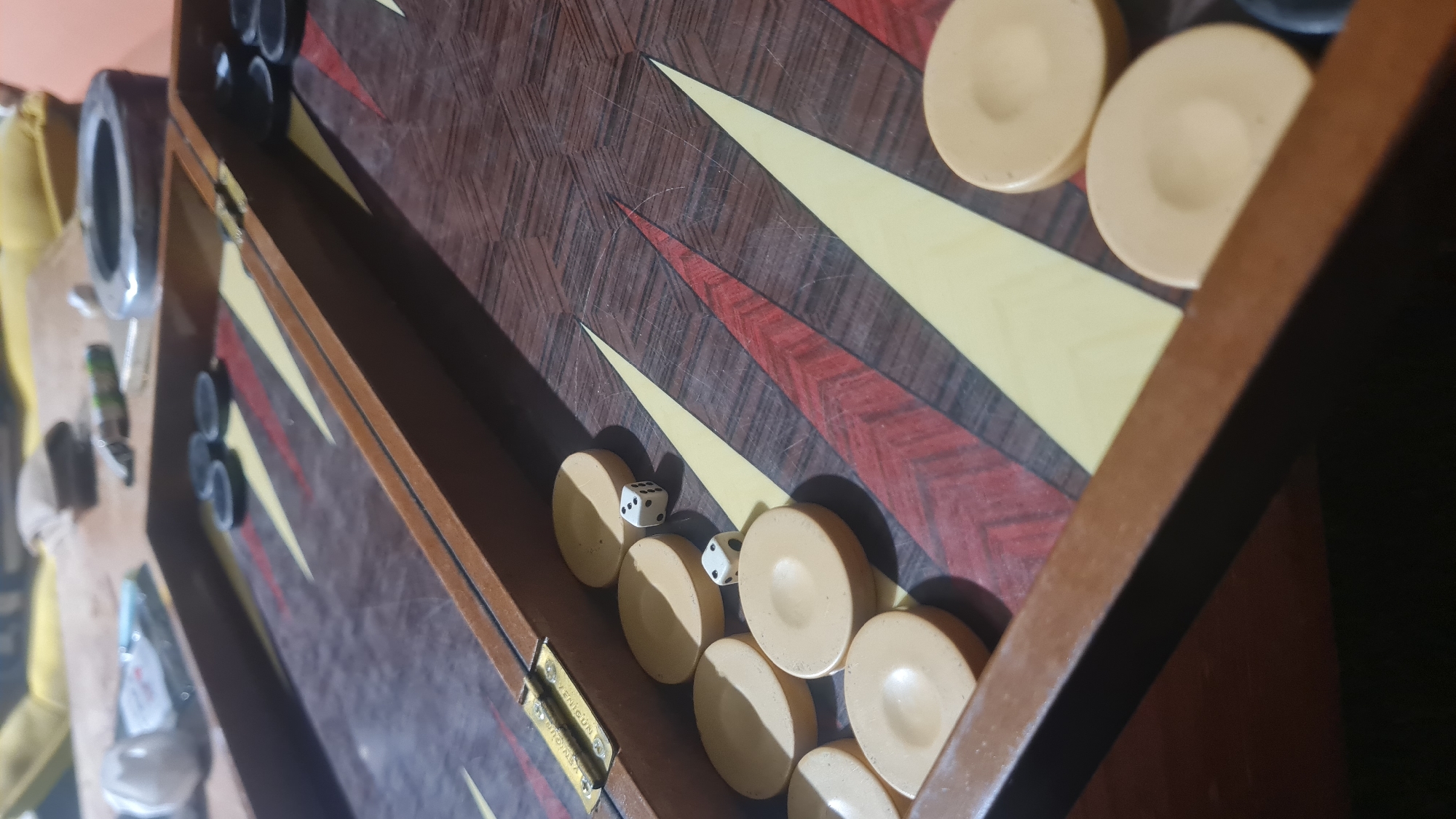 An angled, close-up shot captures a partially open backgammon board. The playing surface is made of dark wood, patterned with alternating yellow and reddish-brown pointed triangles. Clustered on the right side of the board are several cream-colored backgammon checkers, some appearing to be in the bear-off area. A few dark or black checkers are also visible on the opposing side of the board.
Two white dice are present on the board, resting among the cream checkers. One die clearly displays three pips, while the other shows a single pip. The wooden surface of the board exhibits noticeable scratches and wear.
In the out-of-focus background to the left, various indistinct objects can be seen, including a metallic item resembling an ashtray and other household articles, resting on a light-colored surface. Strong shadows are cast by the checkers and the raised edge of the board, suggesting an indoor setting with possibly artificial or directional natural light.
Visible on a brass-colored hinge in the lower left section of the board are engraved words, appearing to be "VERIGIUM" or "VERIGUN" and "ORIGINAL" underneath. The scene depicts a backgammon game setup, likely within a domestic environment in the Lachish Regional Council, Israel.