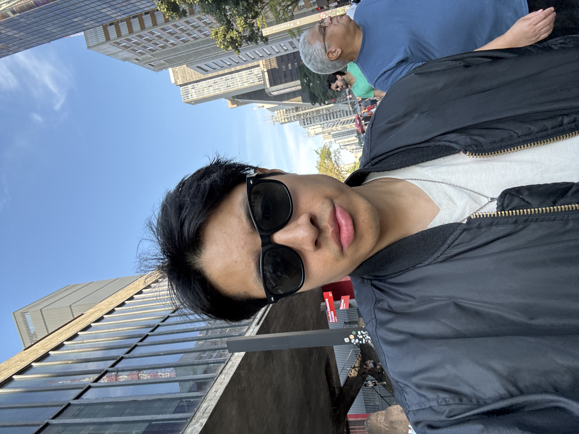 The photograph shows a man taking a selfie. He is wearing sunglasses and a black jacket over a white t-shirt. He is outside in a city, with buildings visible in the background. In the background, there are other people. A street sign in Portuguese reads "R. Pamplona", which indicates the location is likely São Paulo, Brazil. The sky is blue and the weather appears sunny.