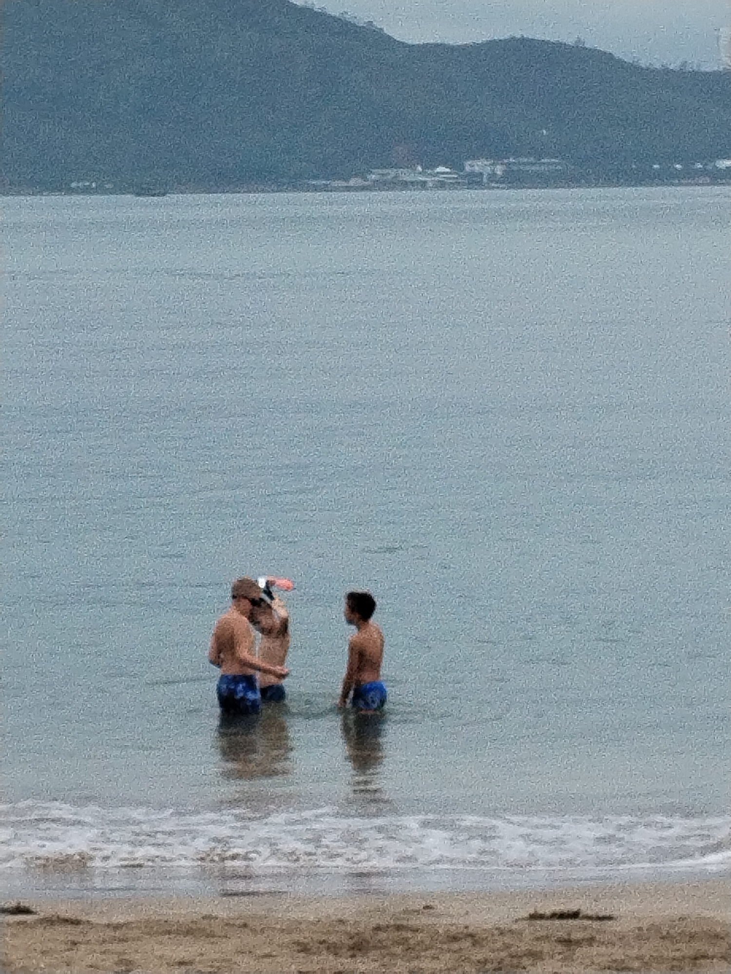 This image depicts an outdoor beach scene on an overcast day in Bac Nha Trang Ward, Vietnam. Three individuals, appearing to be males and likely young adults or teenagers, are standing in the shallow, calm, blue-grey ocean water. They are shirtless and wearing dark-colored swim shorts.
Two of the individuals are positioned closer together in the mid-ground, seemingly engaged in conversation or interaction. The person slightly behind the other appears to be holding a small, pink or orange object, possibly a snorkel mask, a frisbee, or a small float. The third individual stands a short distance to the right of the pair, also partially submerged.
In the immediate foreground, gentle, frothy white waves lap onto a light brown sandy beach. The water exhibits a slightly textured, grainy appearance, which may be due to a combination of lighting conditions and image quality. The background features a distant, hazy mountain range or hilly coastline stretching across the horizon. Faint outlines of buildings or structures can be seen along the base of the mountains by the water's edge.
The sky is overcast, lending a soft, diffused light to the scene and contributing to a muted color palette. No direct sunlight or harsh shadows are visible, suggesting cloudy weather. The overall impression is one of a relaxed moment by the sea. There is no visible text in the image.