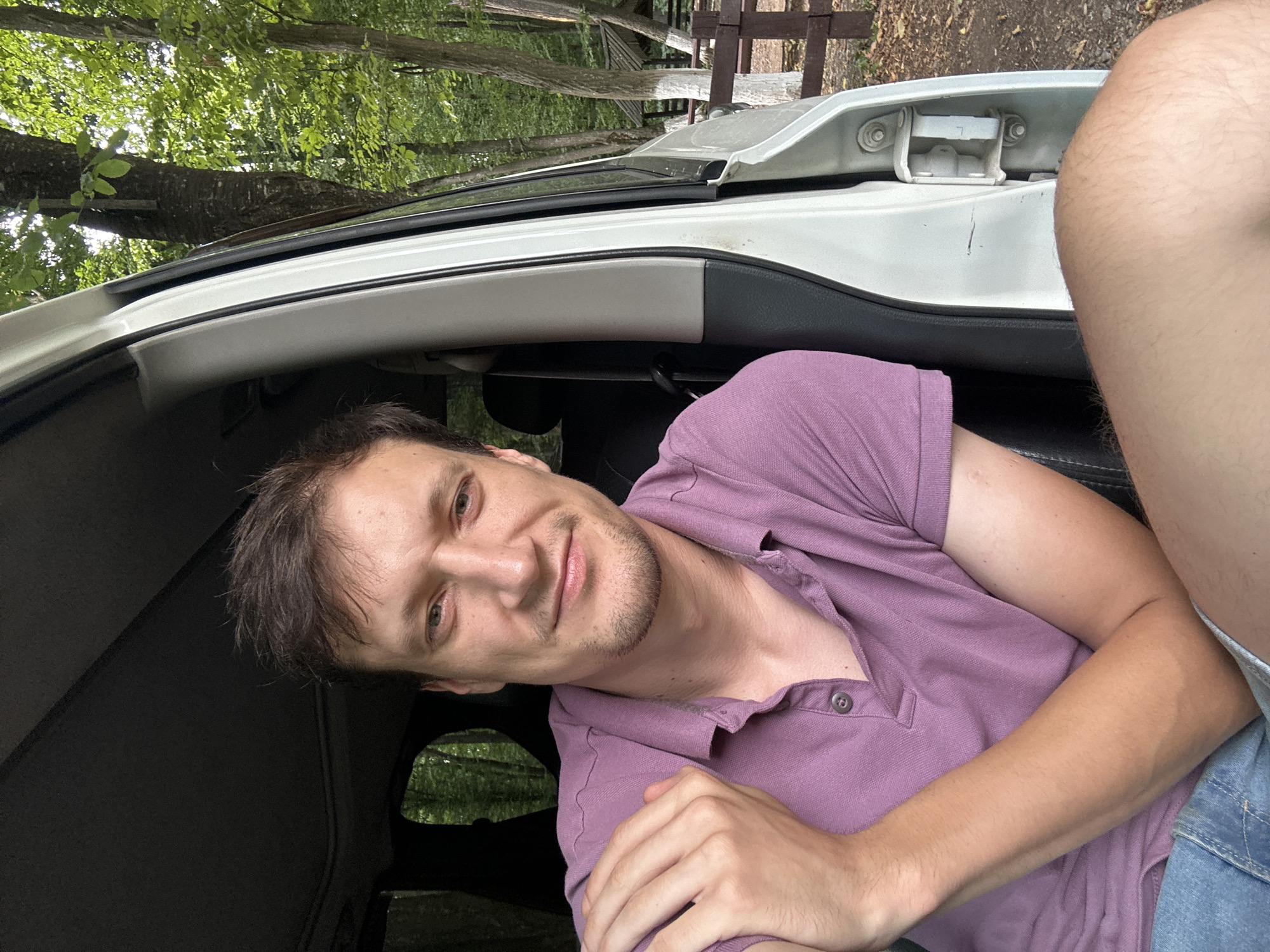 A young adult male with light brown hair and stubble is seated inside a vehicle. He is wearing a light purple polo shirt and denim shorts. His left arm is bent, with his hand resting on his left shoulder. The vehicle appears to be parked outdoors, with trees and a wooden fence visible in the background. The surrounding environment suggests a natural or rural setting, potentially a park or forested area, consistent with the outskirts of Shatava, Ukraine. The lighting indicates it is daytime.