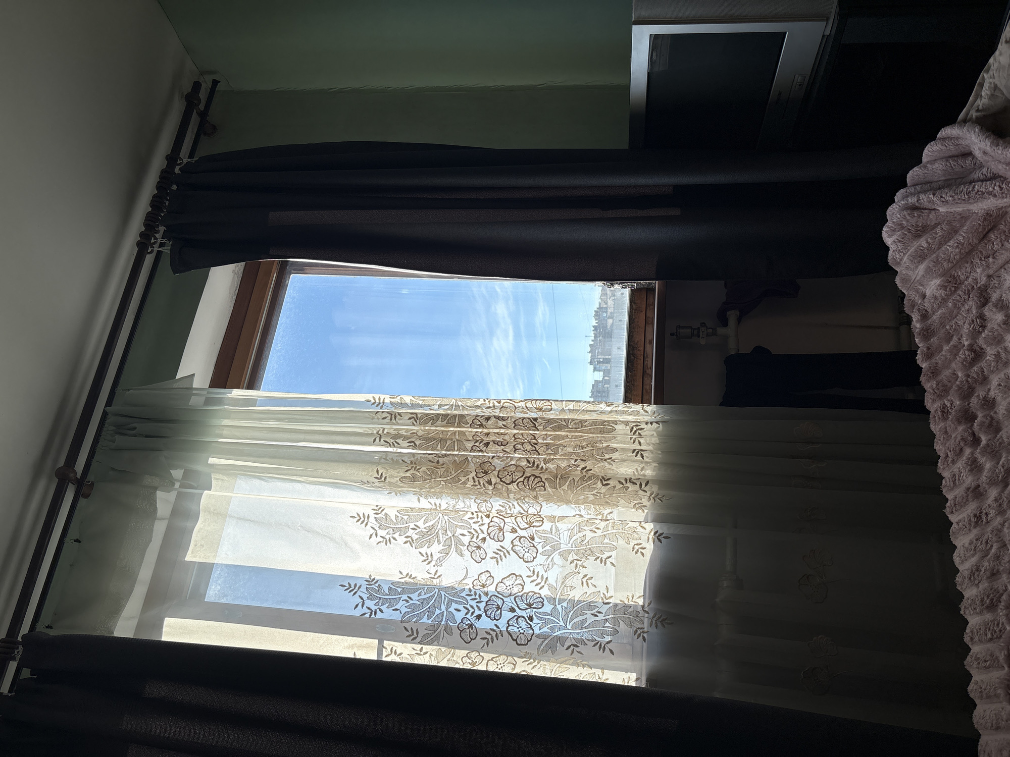 A bedroom scene is captured from a low angle, looking towards a window. Two dark curtains frame the window, one on the left and one on the right, partially obscuring the view. The window is draped with a sheer, patterned curtain in a light beige or cream color, featuring floral designs. Through the sheer curtain, a bright blue sky with scattered clouds is visible, along with a distant cityscape. In the foreground, the edge of a pink, textured blanket or comforter is visible, resting on what appears to be a bed. To the right of the window, a television set is positioned on a dark surface, its screen dark. The walls of the room are painted a muted green color.