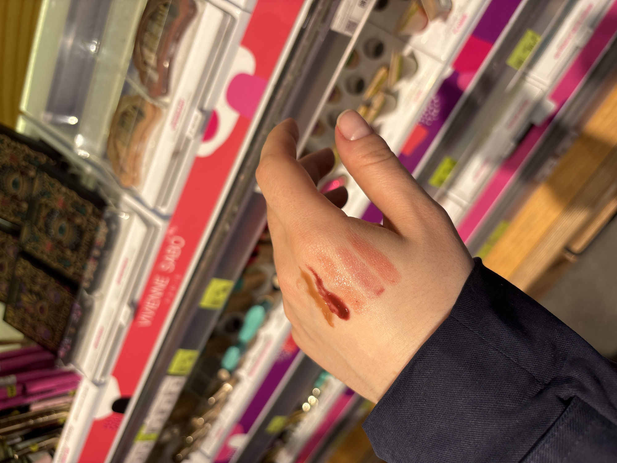 A person is testing makeup swatches on their hand in a store. The person's left hand is in the foreground with three swatches of different shades of makeup. The makeup appears to be lipstick or lip gloss. The store shelves are in the background with various makeup and beauty products. A sign for the brand "VIVIENNE SABO" is partially visible on one of the shelves. Based on the location of the VIVIENNE SABO store, the image was taken in Perm, Russia.