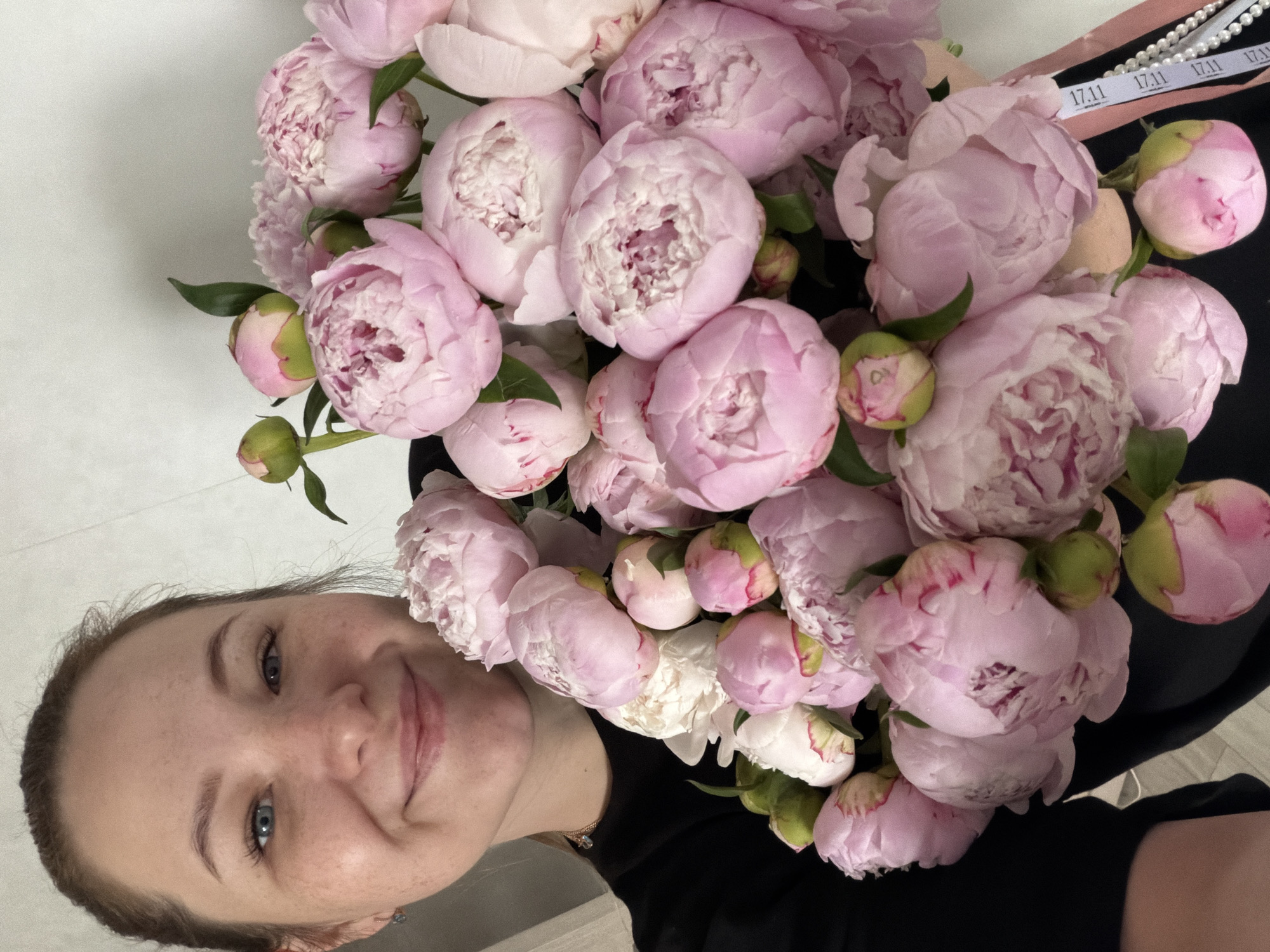A young adult female is captured in a close-up, chest-up portrait, smiling directly at the camera while holding a large bouquet of light pink peonies. Her light brown hair is pulled back, revealing a clear complexion with visible freckles and blue-grey eyes. She wears a black long-sleeved garment, a small stud earring in her left ear, and a thin gold-colored necklace with a small pendant.
The voluminous bouquet of peonies dominates the right side and lower portion of the image, obscuring part of her lower face and torso. The flowers are predominantly light pink, with many in various stages of bloom, displaying densely packed, ruffled petals. Several unopened buds with green calyxes are also visible, along with a few lighter, almost white, blossoms. Tucked into the right side of the bouquet are a white ribbon with repeating black "17.11" text and a strand of white pearls.
The background consists of a plain, light-colored wall, indicating an indoor setting. Soft, even lighting illuminates the scene. A small section of a light-colored floor or surface is visible in the lower-left corner. The specific location within the city of Chaikovsky, Russia, is not discernible from the visual information provided in the image.
