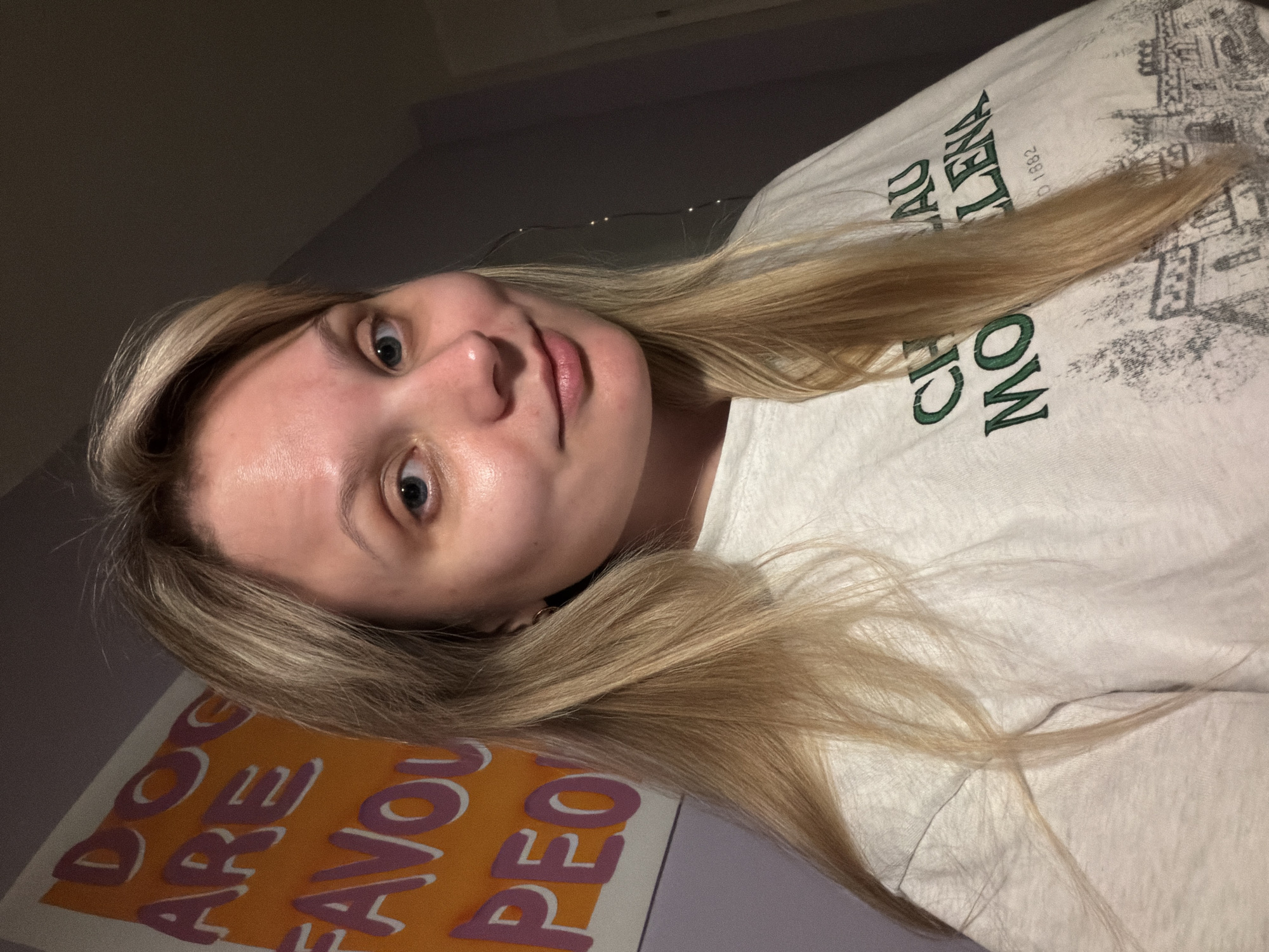 A young woman with blonde hair is facing the camera. She is wearing a white t-shirt with green lettering. In the background, a poster with orange and pink lettering is partially visible. The background wall is light purple. A string of small lights are also in the background. The location is unknown and cannot be determined from the image. Tel-Aviv, Israel, cannot be confirmed or denied.