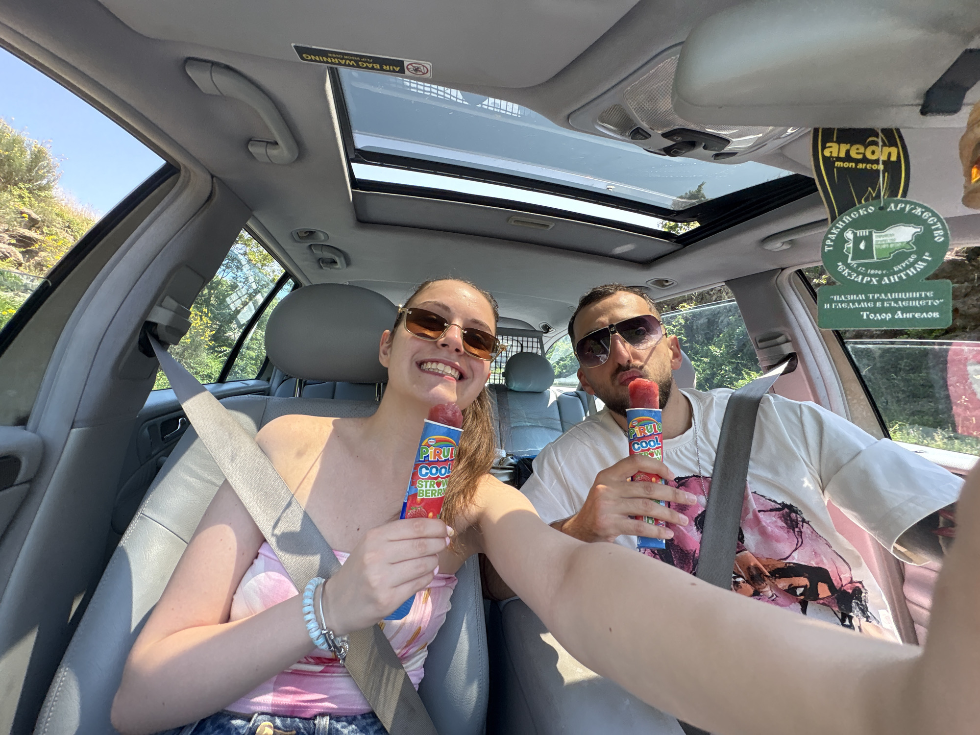 The image displays a selfie taken from inside a vehicle on a sunny day in Tsarevo, Bulgaria, featuring two individuals.
The woman, seated in the front passenger seat on the left, has light brown hair, wears amber-tinted rectangular sunglasses, and a pink and white patterned strapless top. She has multiple light blue coiled and silver-colored charm bracelets on her left wrist. Her right arm is extended towards the camera, holding a "PIRULO COOL STRAWBERRY" popsicle, indicating she is taking the photograph. She is smiling, revealing her teeth.
The man, seated on the right, has a beard and mustache, wears dark aviator sunglasses, and a white short-sleeved t-shirt with a large, multi-colored graphic design in pink, black, and orange. He holds an identical "PIRULO COOL STRAWBERRY" popsicle in his left hand, with his lips slightly puckered. Both individuals are secured by gray seatbelts.
The vehicle's interior features gray upholstery and trim. A sunroof with two panels is visible overhead, along with standard roof-mounted grab handles and control buttons. An upside-down "AIR BAG WARNING FLIP VISOR OVER" label is present on the passenger side sun visor. Two air fresheners hang from the rearview mirror: a yellow, tear-drop shaped one branded "areon mon areon," and a green, circular one with Bulgarian text. The Bulgarian text identifies the green air freshener as from "ТРАКИЙСКО ДРУЖЕСТВО ЕКЗАРХ АНТИМ І" (Thracian Society Exarch Antim I), dated "12.1896 г. - БУРГАС" (12.1896 - BURGAS), and includes the motto "ПАЗИМ ТРАДИЦИИТЕ И ГРАДИМ В БЪДЕЩЕТО" (We preserve traditions and build for the future), attributed to "Тодор Ангелов." A metal mesh partition is visible behind the front seats, separating the passenger compartment from the rear. Through the car windows, a clear blue sky, green trees, foliage, and rocky terrain are visible, indicating an outdoor, natural environment.