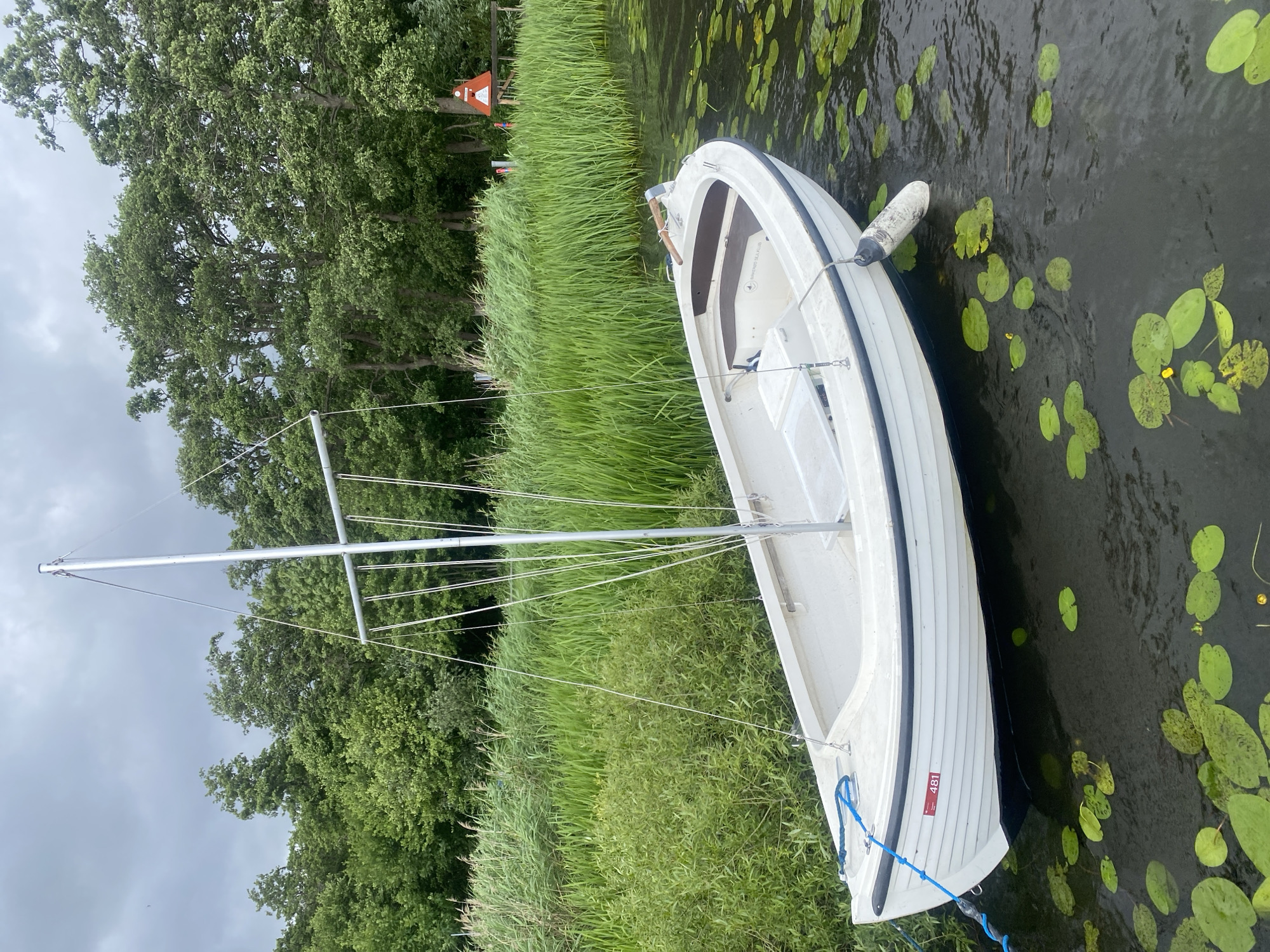 This image depicts a white sailboat moored on a body of water, likely a lake or canal, near a lush green shoreline. The boat, with its mast and rigging, is positioned in the foreground, gently bobbing on the water. Lily pads are scattered across the dark water's surface, with some small yellow flowers visible among them. The background features a dense wall of green trees and reeds, with a cloudy sky overhead. A red and white navigation marker and a wooden structure are visible in the distance on the right side of the frame. The number "481" is affixed to the side of the boat. The scene suggests a serene, natural environment, possibly a park or recreational area. While the prompt mentions Birkerød, Denmark, there are no explicit visual cues in the image to confirm this specific location.