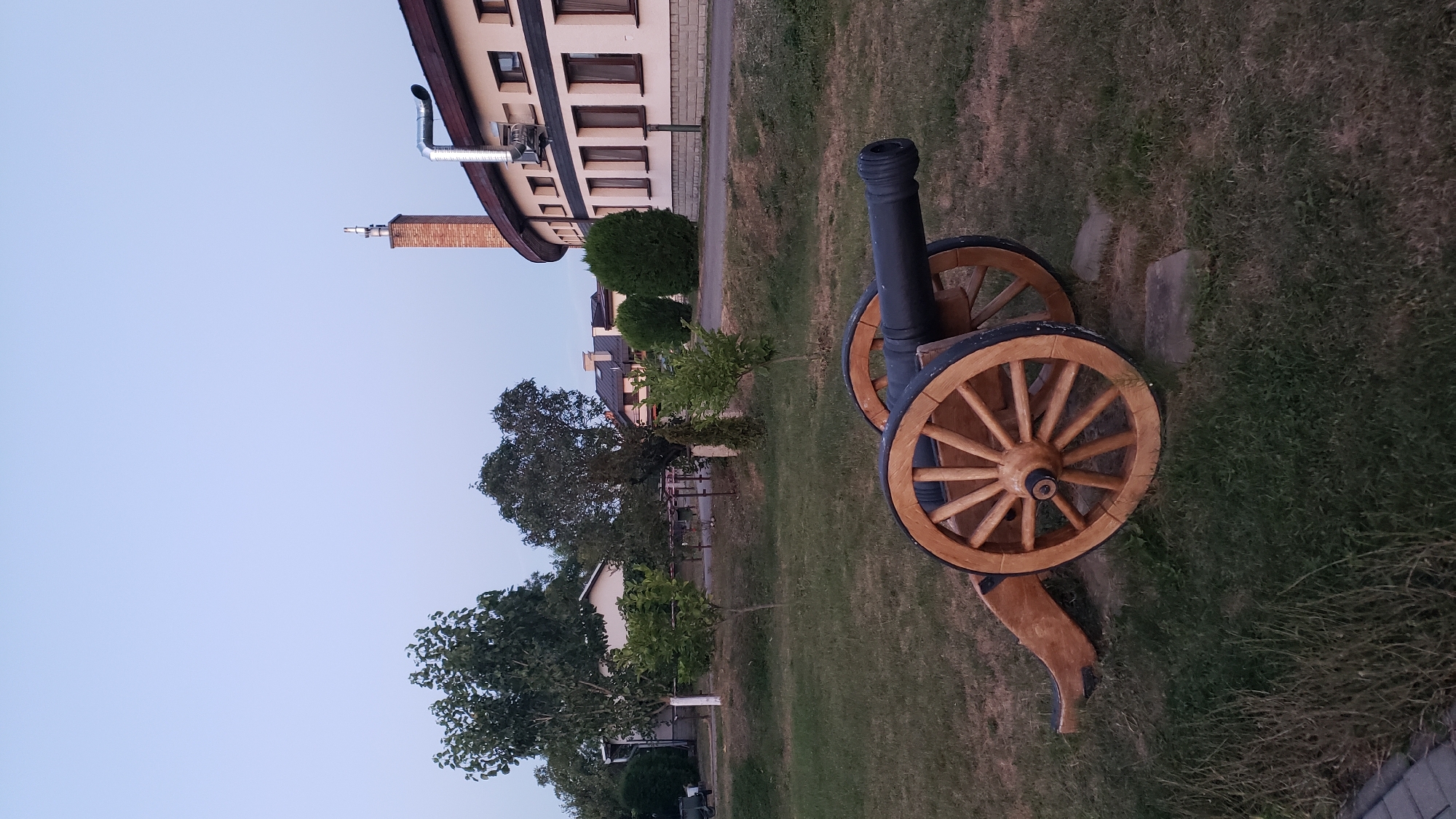 The image shows an antique cannon displayed on a grassy area in front of a curved building. The cannon, made of dark metal and wood, has large spoked wheels. The building behind it is light-colored with a dark roof and many windows. A brick chimney with a metallic vent extends from the roof. In the background, there are trees and smaller houses. The sky is a clear, pale blue, suggesting it is either early morning or late evening. The scene appears to be a public space, possibly a park or a historical site, in the city of Prešov, Slovakia.