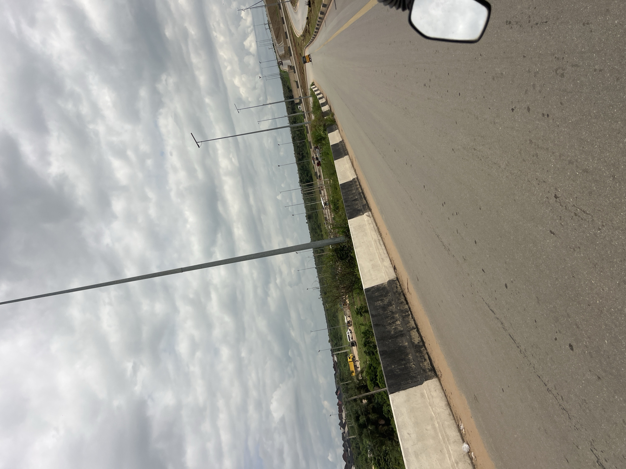 A high-angle, wide shot captures a grey, cloudy sky over a modern highway in Obosi, Nigeria. The highway curves to the right and disappears into the distance. Tall lampposts line the side of the road, spaced evenly apart. On the left side of the highway, there is a concrete barrier with alternating black and white stripes. Beyond the barrier, an area of green vegetation is visible, with some buildings in the far distance on the left. Several vehicles, including a yellow truck, a white van, and a yellow tricycle, are seen on the highway and the adjacent road. The foreground of the image shows a portion of the road and a motorcycle's side mirror reflecting the sky. The overall scene depicts a developing infrastructure in a semi-urban or rural setting under an overcast sky.