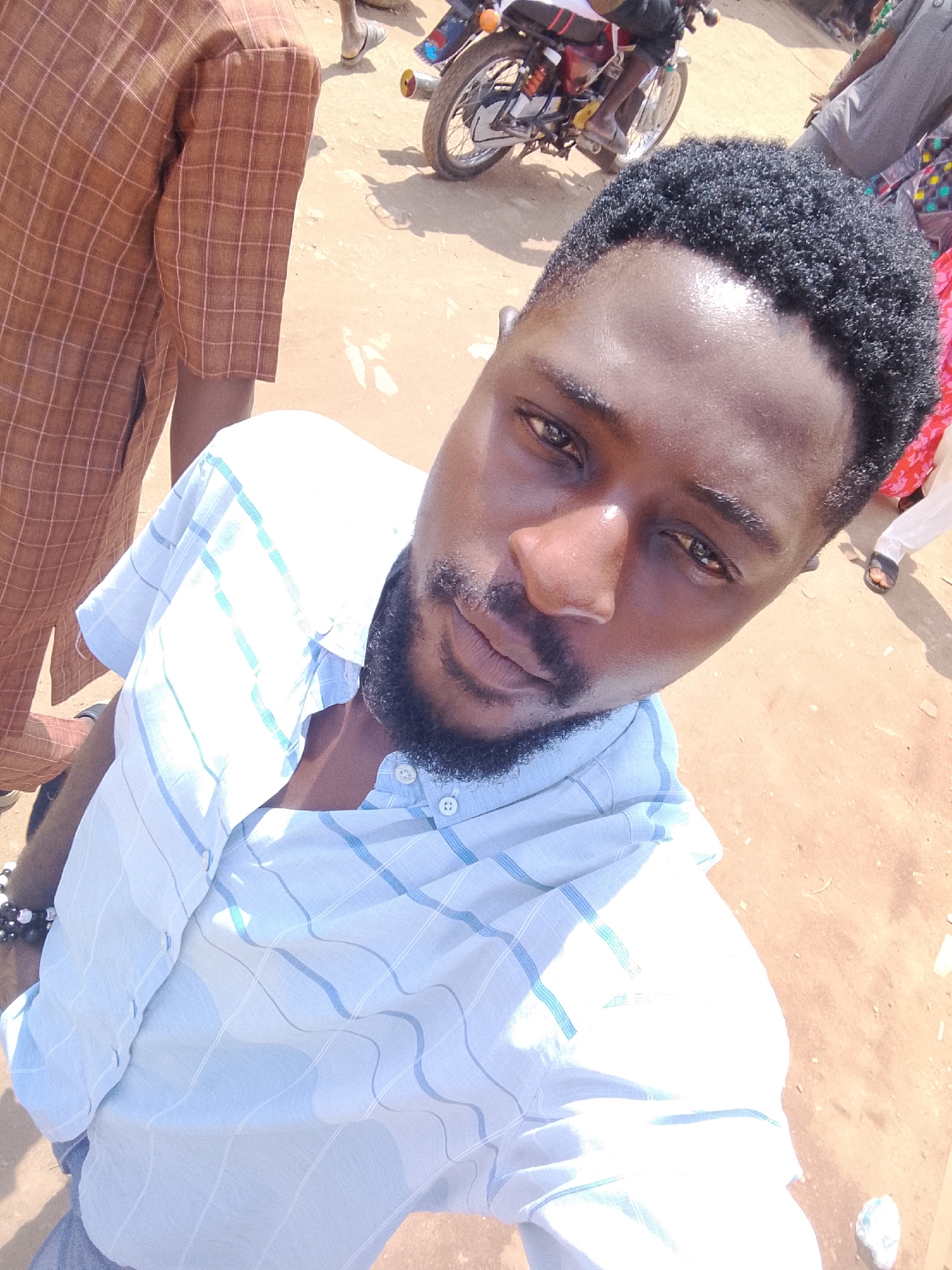 A dark-skinned man with a beard is taking a selfie outdoors in daylight. He's wearing a light-colored, striped button-down shirt. Another person, wearing a brown and tan plaid garment, is partially visible to the left. In the background, there is a motorbike and other people. The ground is a dusty, reddish-brown color. The location appears to be a street scene in Abuja, Nigeria.