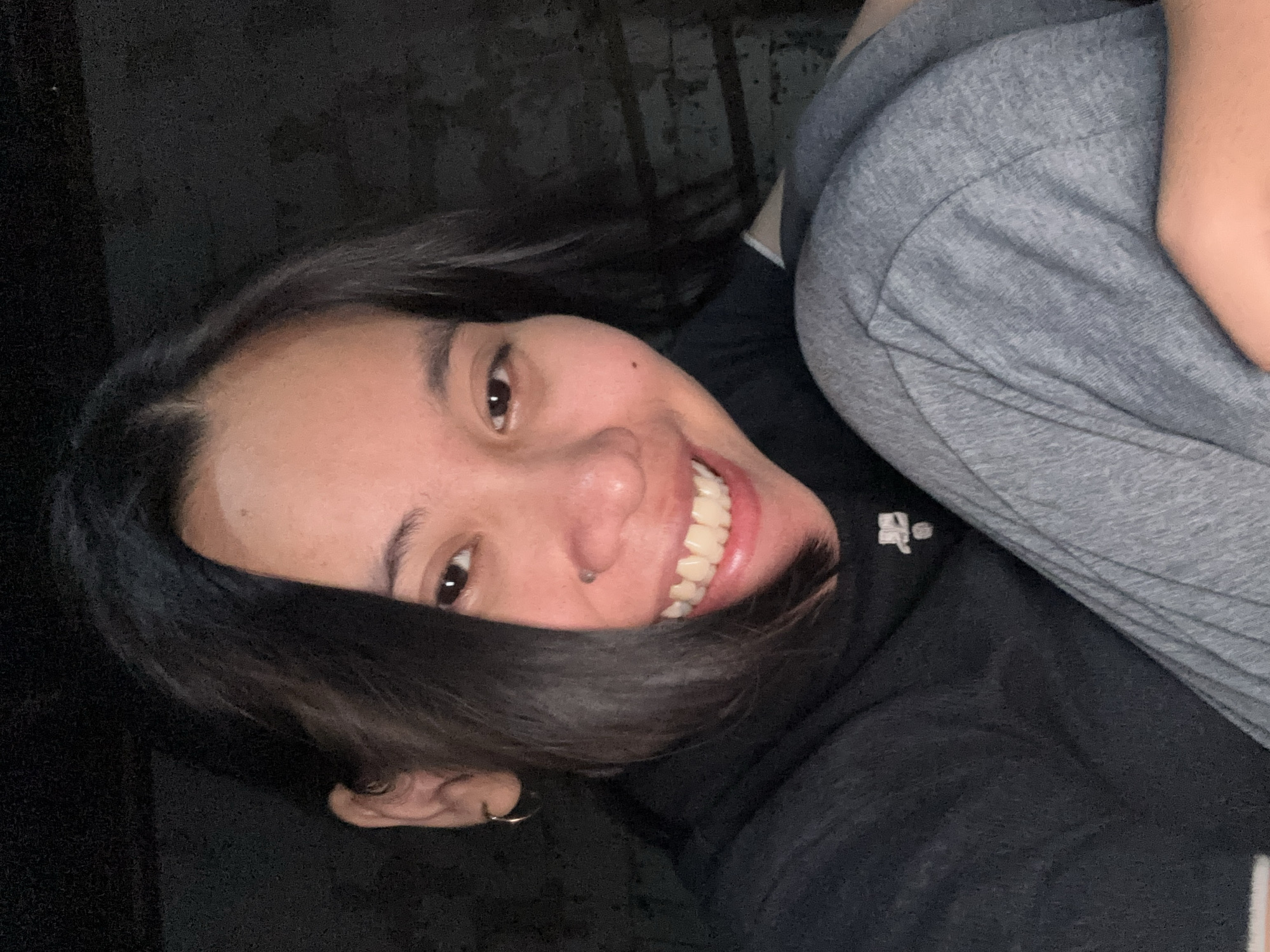 A close-up, eye-level, indoor, selfie-style photograph shows a young East Asian woman with dark hair and brown eyes smiling widely at the camera. She is wearing a black t-shirt with a white logo and grey sweatpants. The background is dark and appears to be a wall with some horizontal lines. The woman is positioned in the lower right quadrant of the frame, with her knees bent and her arms wrapped around them. The lighting is dim, creating a contrast between the woman's face and the dark background. The photograph was taken in East Jakarta, Indonesia.