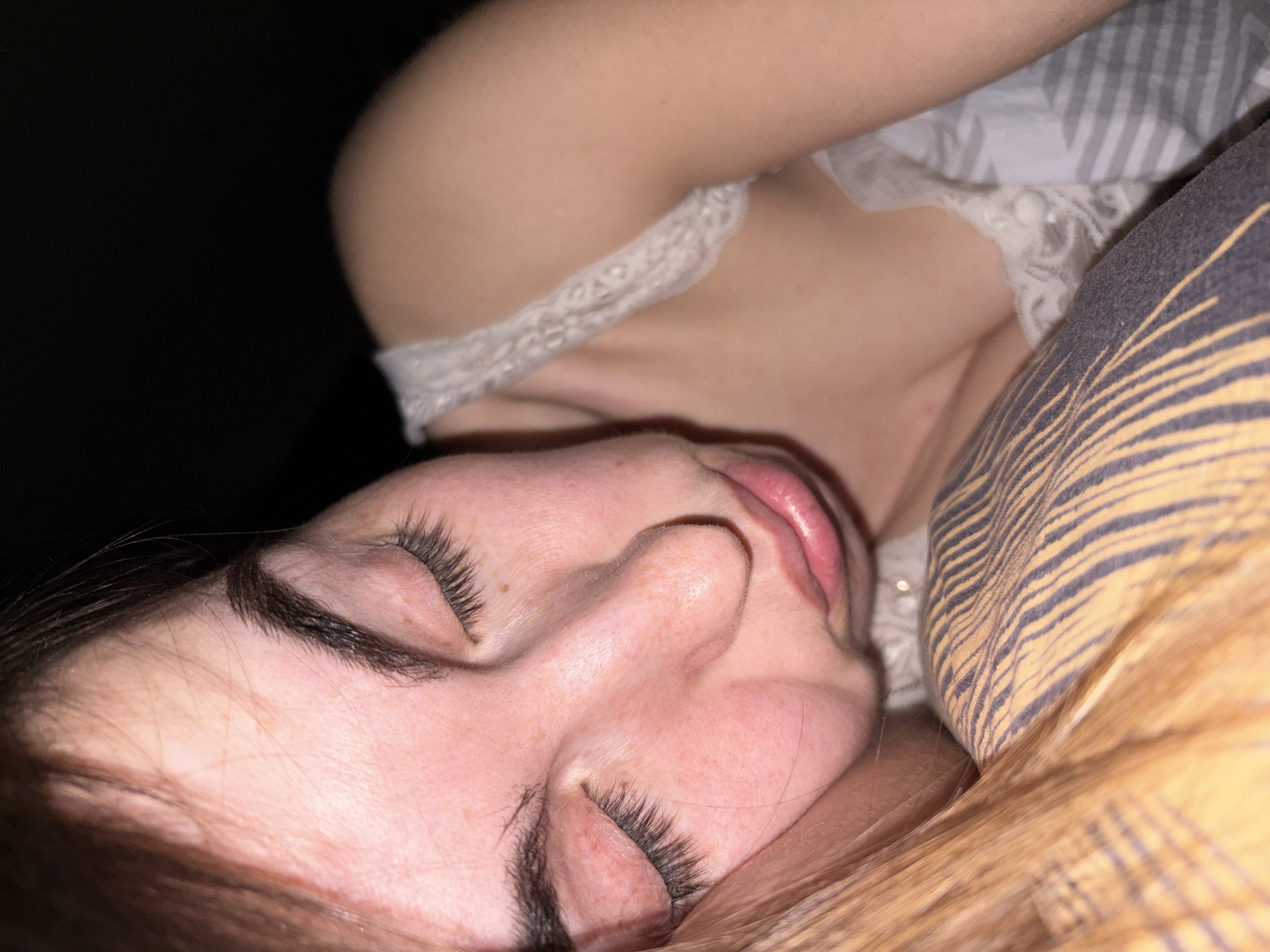A close-up, high-angle shot depicts a fair-skinned woman asleep on a pillow. The woman is lying on her right side. Her eyes are closed, and her long eyelashes are visible. She has dark eyebrows and light-colored hair at the bottom of the frame, near her face. Her skin is pinkish, and her lips are slightly parted. She appears to be wearing a light-colored top with lace detailing around the neckline. The pillow has a striped pattern in shades of yellow and gray. The background is dark and underexposed, making it difficult to discern details. There is no indication of the location being Izhevsk, Russia.
