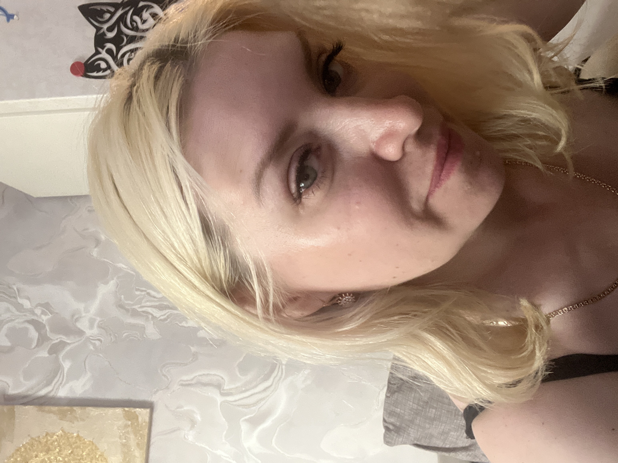 An upward-angle selfie captures a fair-skinned blonde woman with light blue eyes and light pink lipstick. She has on a black tank top and a thin gold chain necklace. Her right earlobe is adorned with a small, sparkling flower-shaped earring. The background shows a textured wall with a marbled pattern. To the upper left, a square artwork with a gold sunburst design is visible. To the upper right, a black silhouette of a wolf's head with a red dot on its forehead is affixed to the wall. The woman is positioned in front of a light gray sofa. The overall setting suggests an indoor residential environment. The image was captured in Tyumen, Russia.