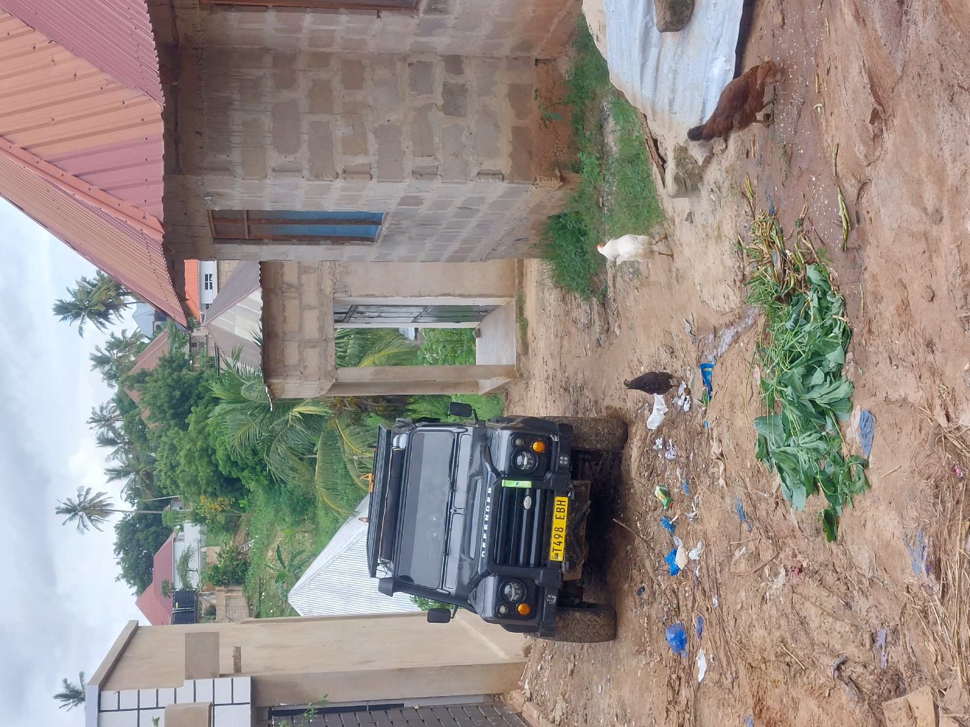 The image depicts a black Land Rover Defender parked on a dirt road in what appears to be a residential area. Chickens are visible near the vehicle. The environment includes unfinished brick buildings, corrugated metal roofs, palm trees, and litter on the ground. The location is most likely Dar es-Salaam, Tanzania, as indicated by the license plate "T498 EBH" which is a Tanzanian license plate format.