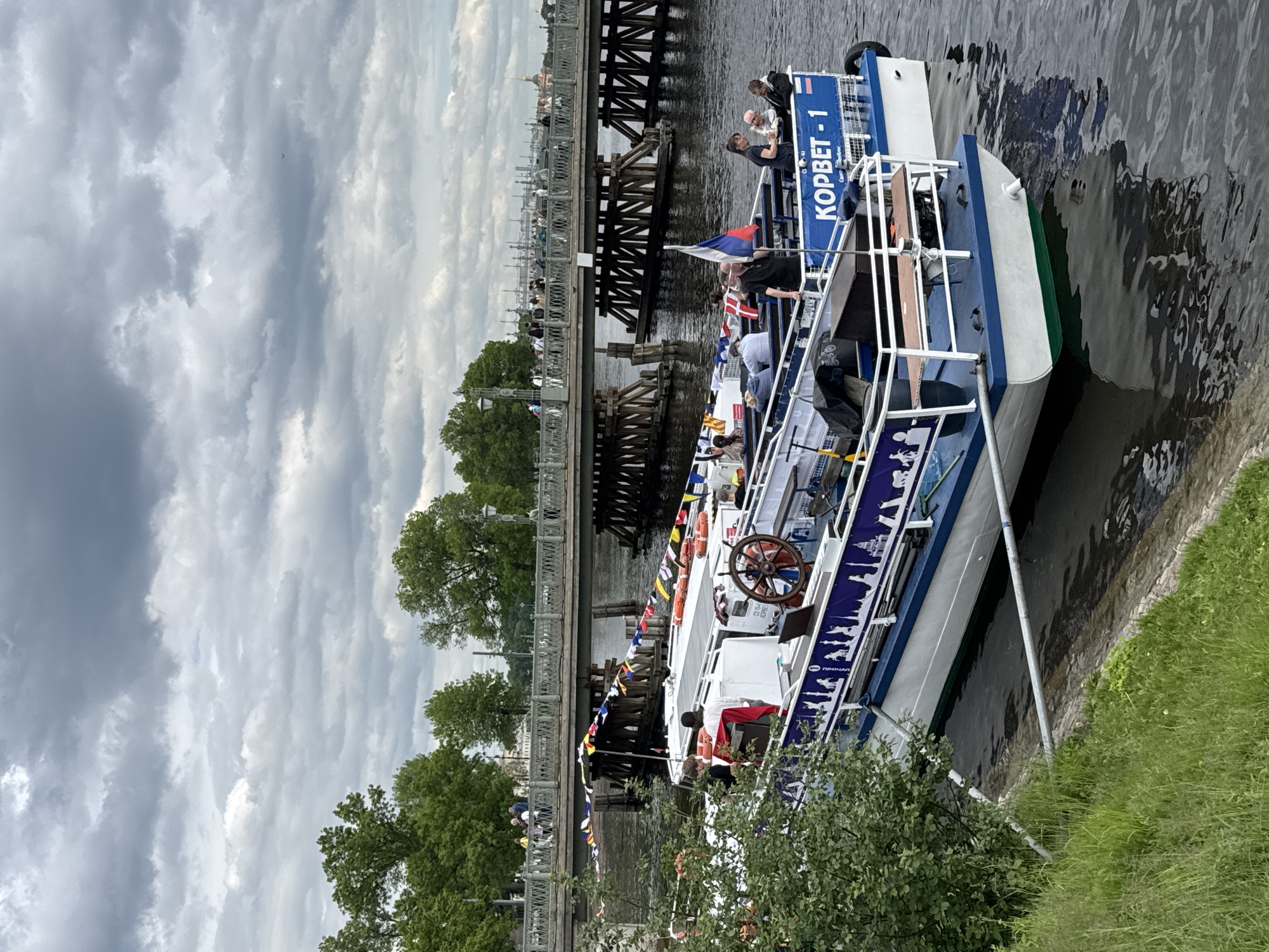 The image captures a sightseeing boat, named "КОРВЕТ-1" (KORVET-1), on a waterway in Saint Petersburg, Russia, under an overcast daytime sky. The boat, painted white and blue with a green stripe at the waterline, is seen from a slightly elevated perspective, seemingly docked or moving slowly near a grassy bank on the right.
On board, several people, including passengers and potentially crew, are visible. Some are standing, observing the surroundings, while one individual is positioned near a large wooden steering wheel, suggesting they are at the helm. The boat is decorated with various flags, including the Russian national flag and a Danish flag, along with strings of colorful signal flags. Lifebuoys hang from the railings, which feature a white silhouette of cityscapes.
In the background, a substantial bridge spans the waterway, with both wooden structural elements and a metal pedestrian railing. Several people are visible walking across the bridge. Beyond the bridge, an urban landscape unfolds, featuring green trees and distant city buildings, including a faint golden spire characteristic of Saint Petersburg's architecture. The sky is largely covered by dense, grey clouds, indicating an overcast day, but the scene is well-lit. The water in the foreground reflects the cloudy sky and the boat's hull, showing gentle ripples. The text "КОРВЕТ-1" is clearly visible on the boat's side, along with smaller text that includes "ПРИЧАЛ" (Pier/Dock) and other less legible Russian words.