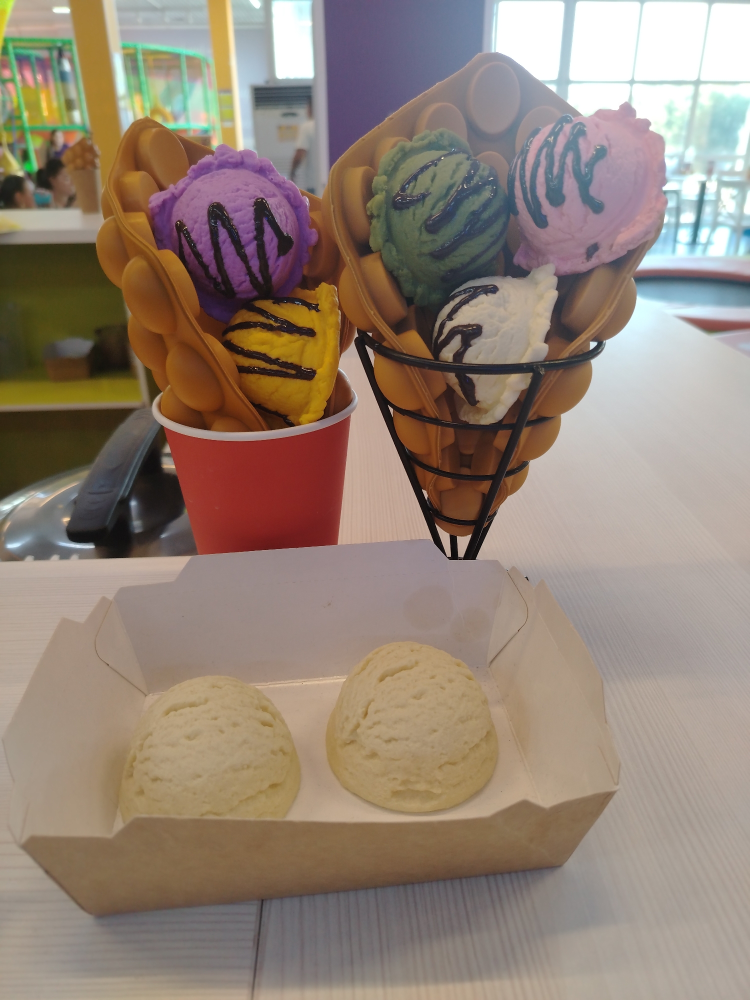 A close-up shot of ice cream cones and scoops is presented, likely on a white table. In the foreground, two scoops of light-colored ice cream are nestled in a white and brown paper container. Behind them, two waffle cone desserts are arranged. One cone, held in a red cup, features scoops of purple and yellow ice cream, both drizzled with dark chocolate sauce. The other cone, supported by a black wire stand, contains scoops of green, pink, and white ice cream, also with chocolate drizzle. The background appears to be an indoor play area, with colorful structures and blurred figures of people visible. The overall lighting suggests an indoor setting.