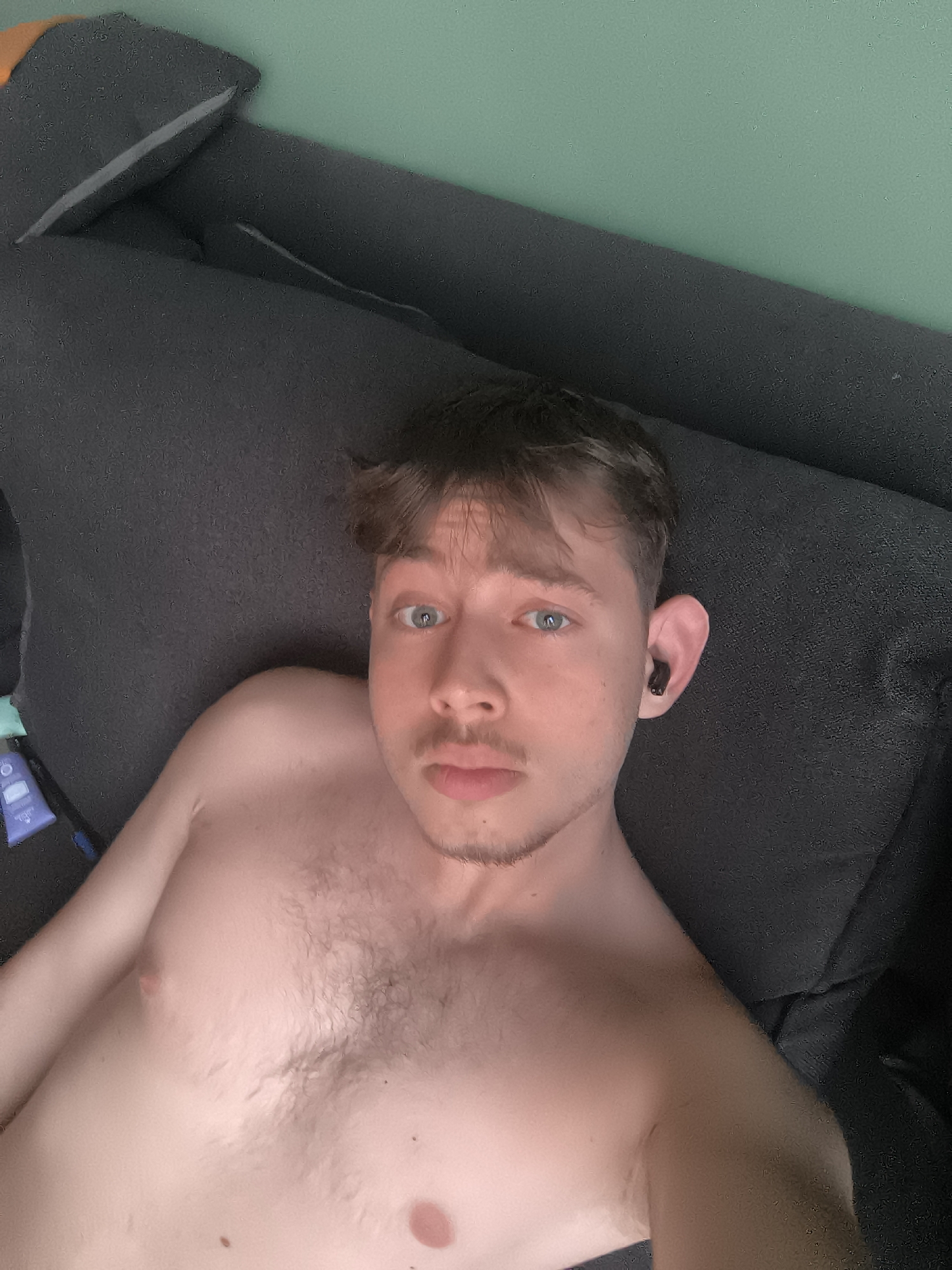 A shirtless, fair-skinned male with brown hair, blue eyes, and stubble is lying on a dark gray sofa. He has a black wireless earbud in his right ear. The background consists of a light green wall. There is a tube of lotion and a blue object on the sofa to the left. The location cannot be determined from the image.