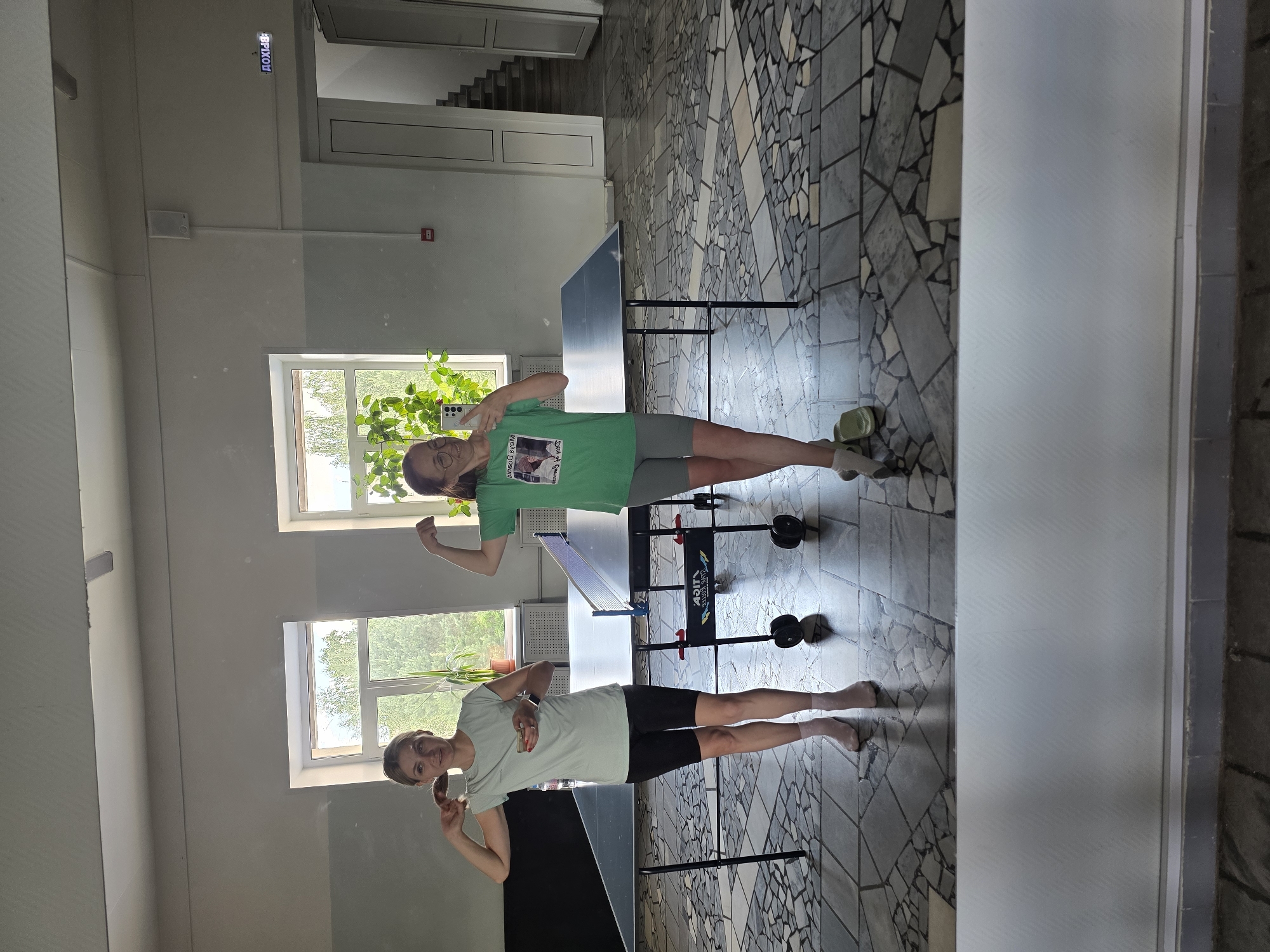 The image shows a reflection in a mirror of two women in a room. One woman is wearing a green t-shirt and grey shorts, and she is posing with her fist clenched. The other woman is wearing a light blue t-shirt and black shorts, and she is making a peace sign with her fingers. Both women appear to be smiling and have a playful demeanor.
They are standing in a room with tiled floors and large windows that look out onto trees. There is a table tennis table between them. The room appears to be well-lit, suggesting it is daytime. The setting seems to be an indoor recreation area or gymnasium. There is text visible in the reflection: "ДОХОД" (meaning "income" or "revenue" in Russian) is written on a blue sign above a doorway. The overall impression is one of a casual and fun moment captured in a mirror.
