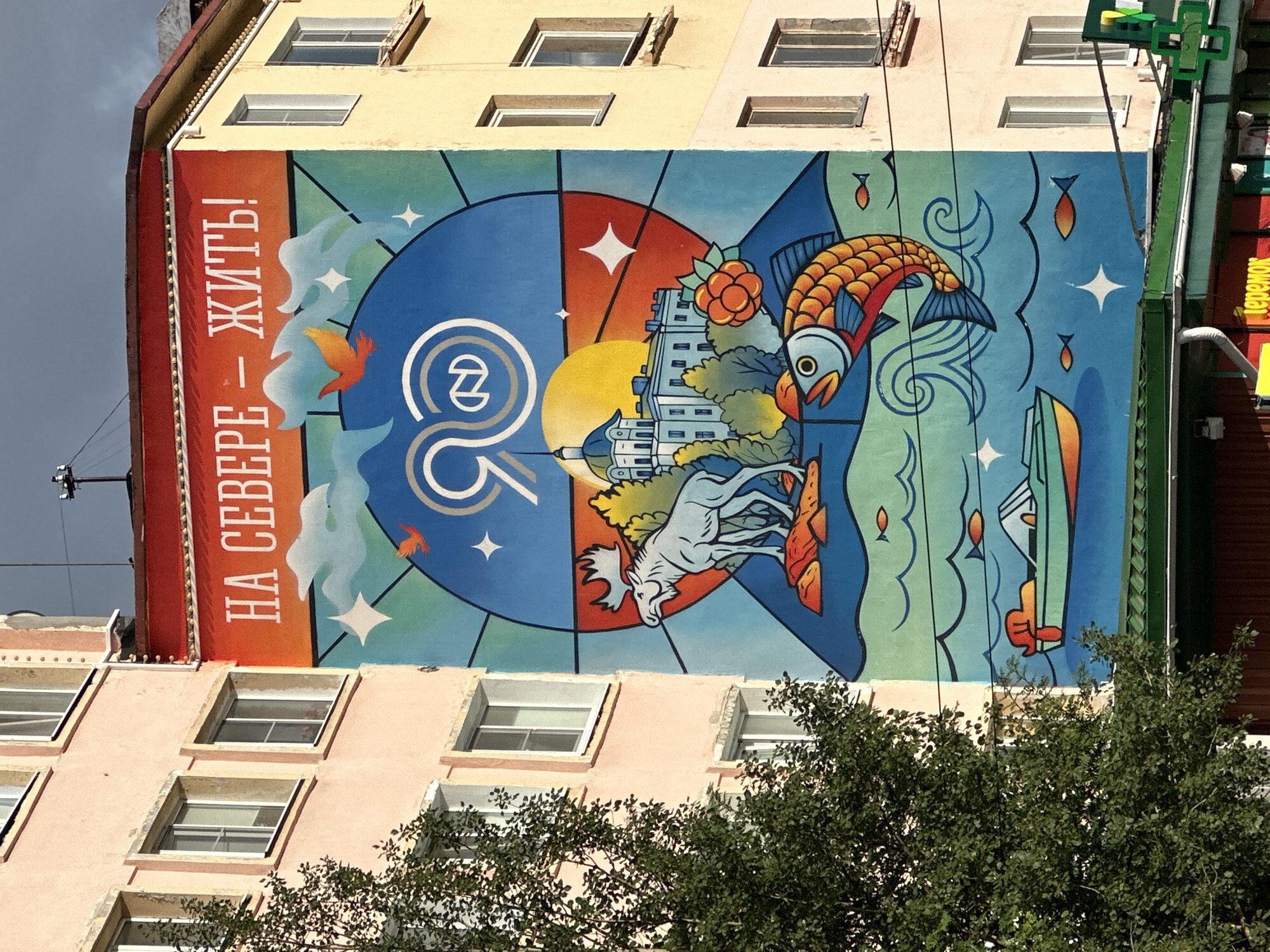 This is a photograph of a mural painted on the side of a residential building in Monchegorsk, Russia. The mural is a colorful, stylized depiction of northern life. The top of the mural features text in Russian that reads "На Севере - Жить!" which translates to "To Live in the North!". Below the text, a large blue circle dominates the center, with a stylized "90" in white and a stylized "N" inside a circle at its center. This likely commemorates an anniversary.
The mural contains various elements representative of the north: a white moose stands on a rocky landscape, with a domed building in the background. A large, orange fish with blue fins swims in a stylized blue wave. A small boat with an outboard motor is depicted on the water, and several small fish are scattered throughout the scene. There are also stars and clouds painted in the sky. A single orange cloud-like shape with bird-like features flies in the upper right quadrant.
The building itself is a multi-story structure with light pink and yellow facades, featuring numerous windows. A large, leafy tree is partially obscuring the left side of the building and mural. The sky above is overcast with a dark, cloudy appearance. The bottom of the image shows the eaves of the building and the facade of businesses below, including a green cross symbol which indicates a pharmacy. The overall impression is a vibrant artistic representation of the unique environment and spirit of life in the northern region of Russia.