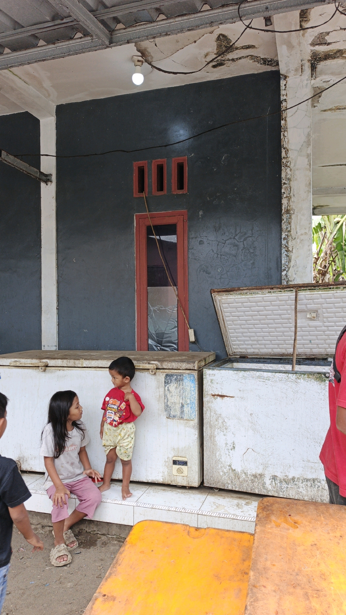 In Cibenda, Indonesia, two young children, a girl and a boy, are standing in front of a dark gray building. The girl, seated on the floor with her legs crossed, wears a light gray t-shirt and pink pants. She looks towards the boy. The boy, standing, is dressed in a red t-shirt with a graphic print and yellow patterned shorts. He is facing the girl, with his right arm extended.
Behind them, a red-framed door with three small rectangular windows is visible on the building's facade. The door appears to have a broken window. In front of the building, two large white chest freezers are positioned side-by-side. The left freezer is closed, while the right freezer is open, revealing its white interior. The area in front of the freezers is tiled.
To the left of the scene, the back of another person, wearing a black t-shirt and blue jeans, is partially visible. To the right, the upper body of another person in a red shirt is also visible. In the foreground, two bright yellow rectangular objects, possibly tables or benches, are present.
The building's exterior shows signs of disrepair, with peeling paint and visible cracks on the ceiling and a pillar. Overhead, a metal roof structure is apparent. A white light fixture hangs from the ceiling. Electrical wires are strung across the scene. The ground in front of the building appears to be dirt or gravel.