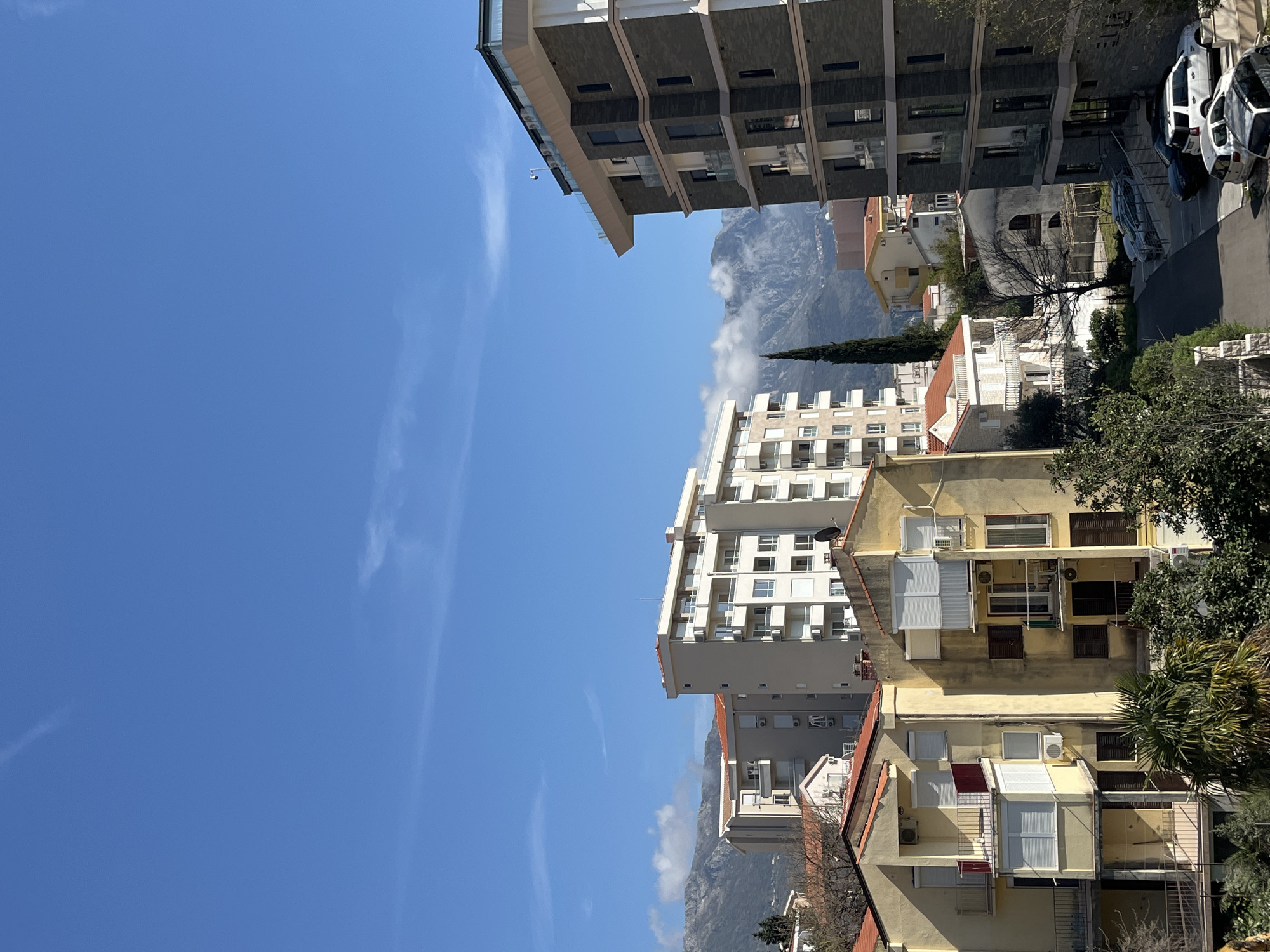 The image shows a view in the city of Boreti, Montenegro, featuring a bright blue sky with wispy clouds above a landscape of buildings and a mountain range. The architecture includes a mix of multi-story residential buildings with various colors and designs. Some buildings have balconies, air conditioning units, and window awnings. The mountains in the background are partially shrouded in clouds. The foreground contains trees and vegetation, obscuring the view of the street below. Parked cars are visible on the street.