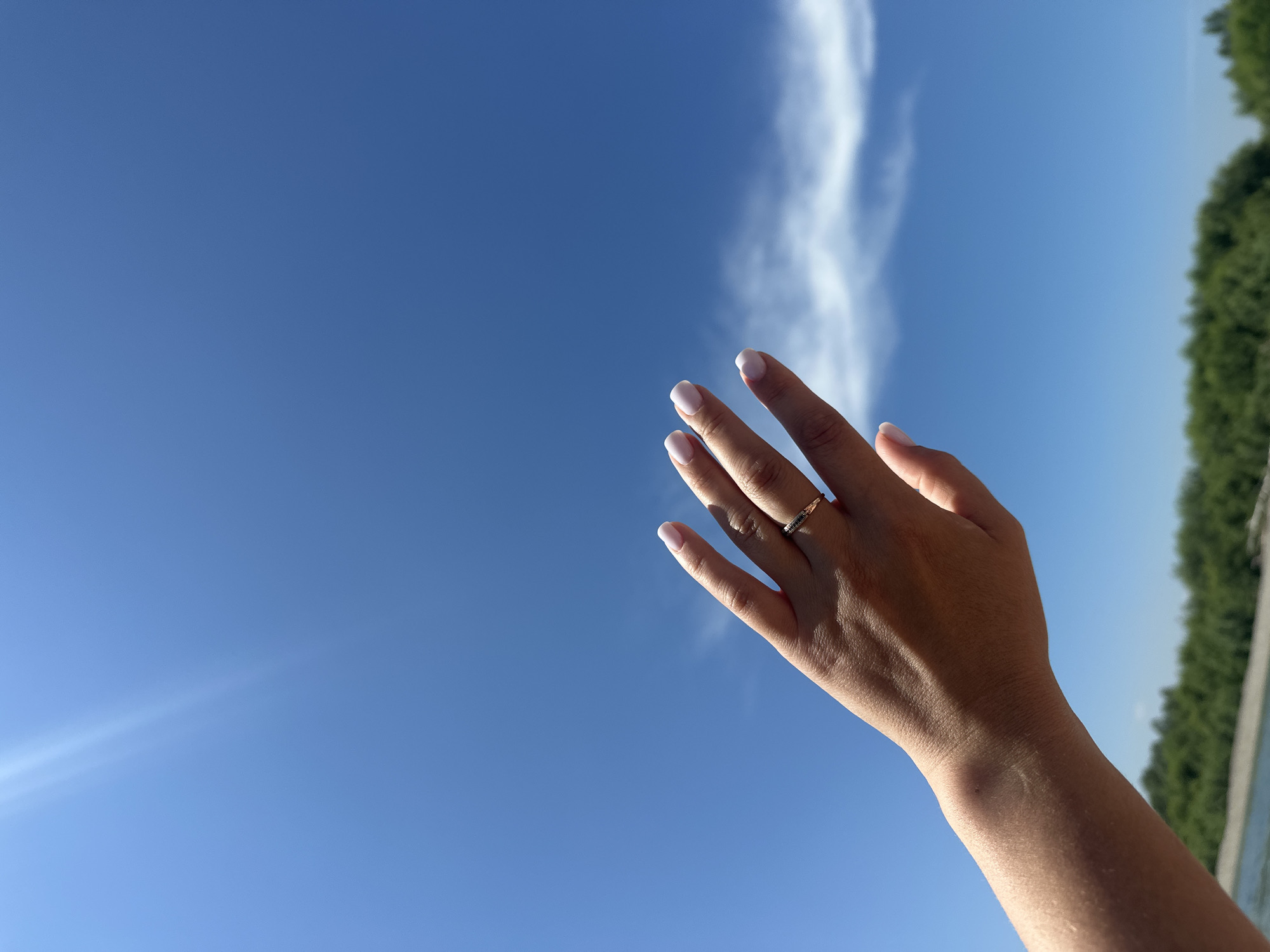 A close-up, slightly low-angle shot captures a person's left hand and forearm against a bright blue sky. The hand is extended upwards and slightly to the right, with fingers splayed. The fingernails are painted with white polish. A thin, gold-colored ring with small dark stones is visible on the ring finger. The skin of the forearm and hand appears tanned. Sunlight flares in the upper left corner of the frame. In the background, a blurred line of green trees and a body of water, possibly a river, are visible, suggesting an outdoor setting. The image context implies this scene is located in Bely Yar, Russia.