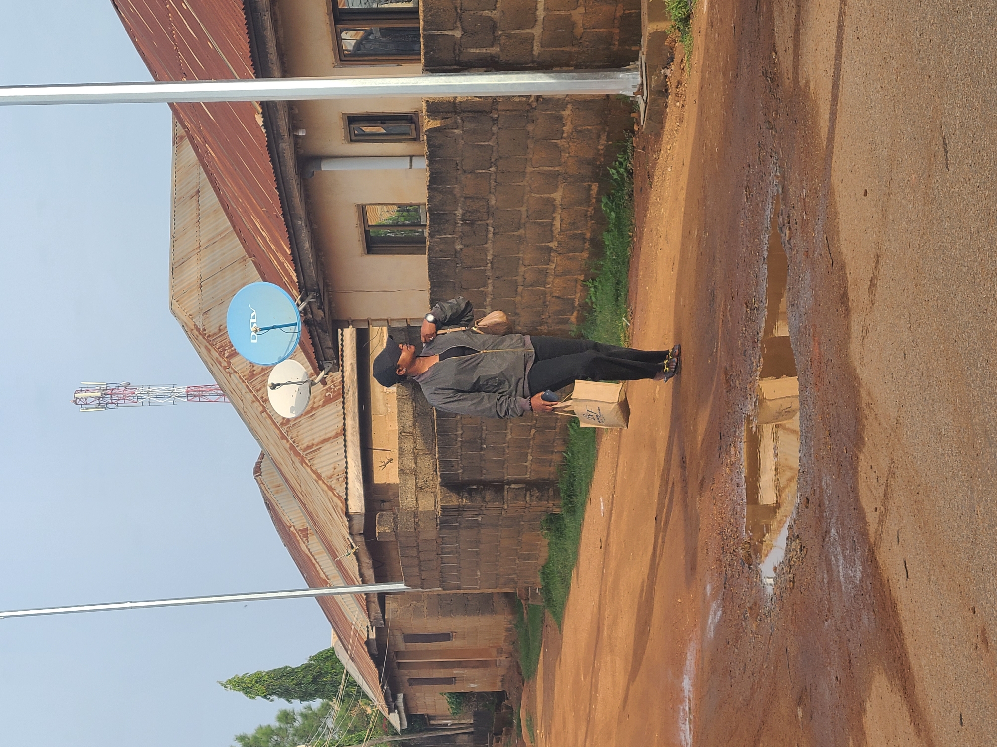 The image shows a man walking on a dirt road in what appears to be a residential area in Jos, Nigeria. The road has a large puddle. The man is wearing a jacket, black pants, and a hat and carrying a brown paper bag. Behind the man, there is a building with a rusty corrugated iron roof and a brick wall. A blue satellite dish is attached to the roof. In the background, there is a communications tower. Metal poles, likely for street lighting, are visible on the left side of the image. The image is taken during daylight hours.