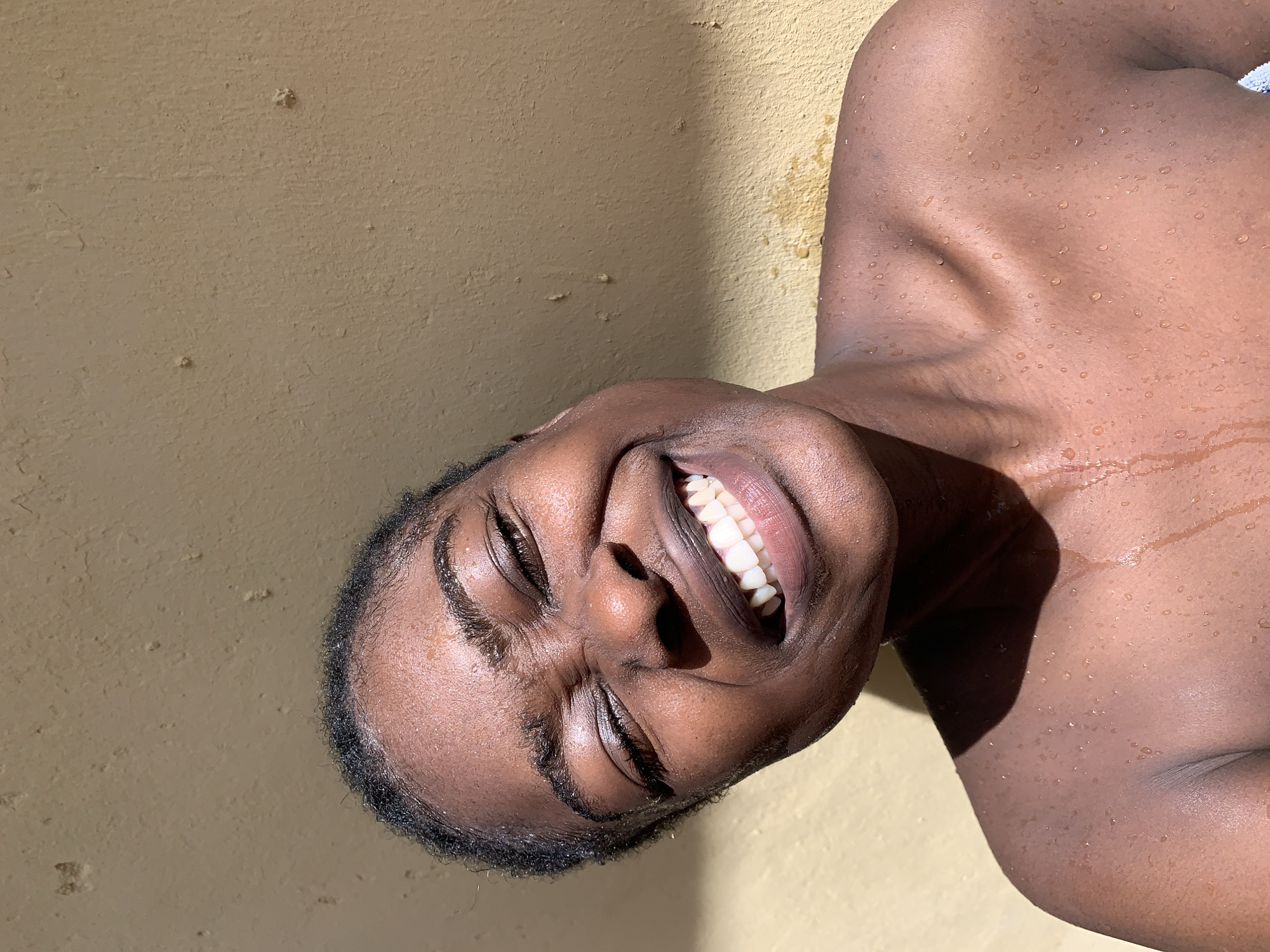 A dark-skinned person with wet skin, cropped at the chest, is positioned at an angle against a yellow wall. The person is smiling with closed eyes, facing towards the viewer. Water droplets are visible on the person's skin. The background appears to be an exterior wall. There is no information to confirm this photo was taken in Rafin Par, Nigeria.
