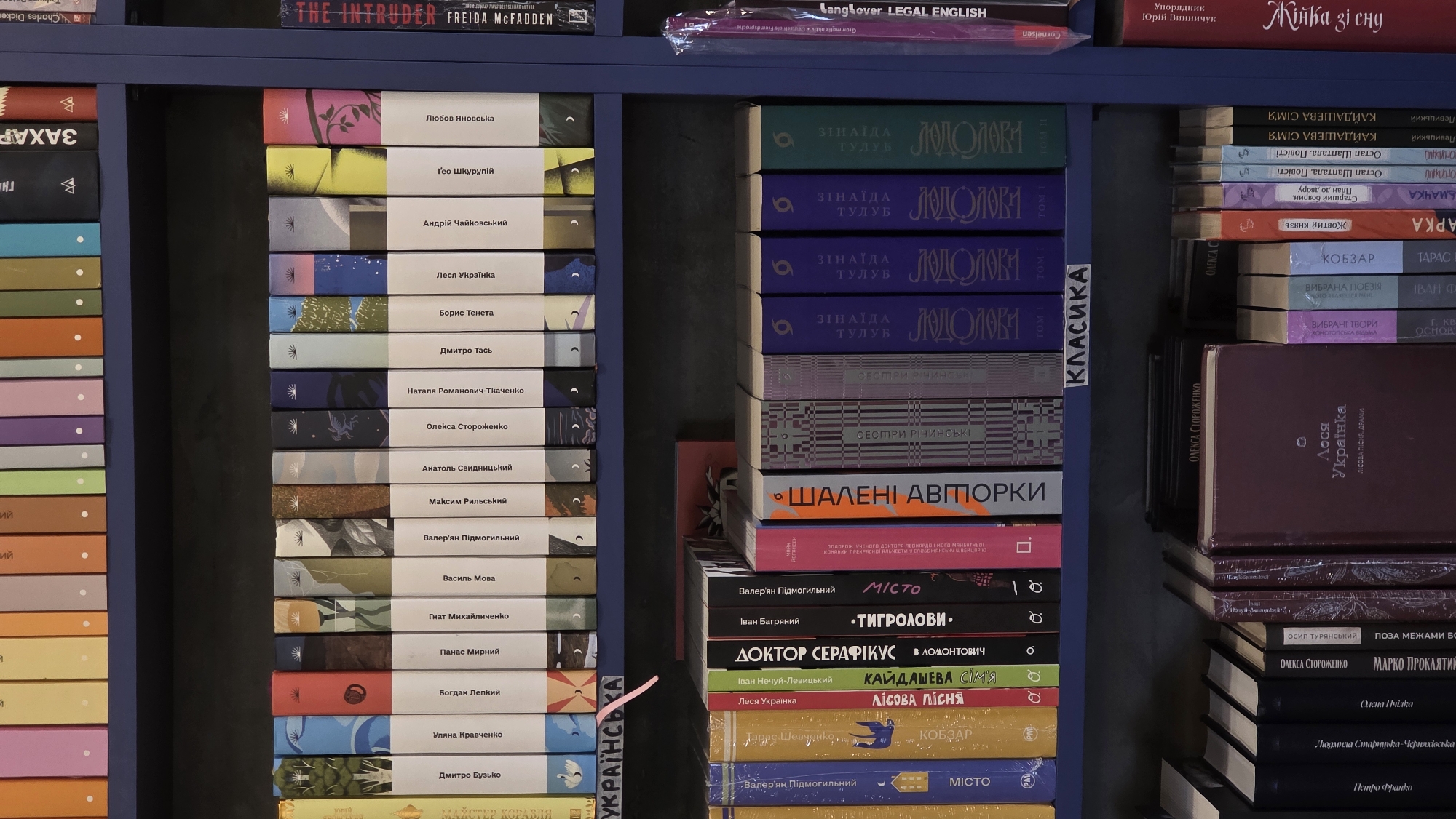 The image shows a close-up, straight-on view of bookshelves filled with books. The shelves are dark blue. The books are predominantly hardcover and paperback, with spines in various colors and designs, many featuring text in Cyrillic. Some spines are plain white or cream, while others have intricate illustrations or patterns. There are also some books with plastic wrappings.
The books are arranged in several rows, with the top row displaying spines of different colors, possibly from a distance. The second and third rows from the top are packed with books whose spines are clearly visible. A small pink sticky note with Cyrillic writing is attached to the edge of the second shelf from the top, on the left side. The bottom rows show more books, some with prominent titles in Cyrillic. One large, maroon-colored book with Cyrillic text is visible on the bottom shelf.
There are no people or visible actions taking place. The environment appears to be an indoor space, likely a bookstore or library, given the extensive collection of books. The lighting is even, suggesting ambient indoor lighting.
Without additional information or context, it is not possible to determine the specific location within the city of Kyiv, Ukraine.